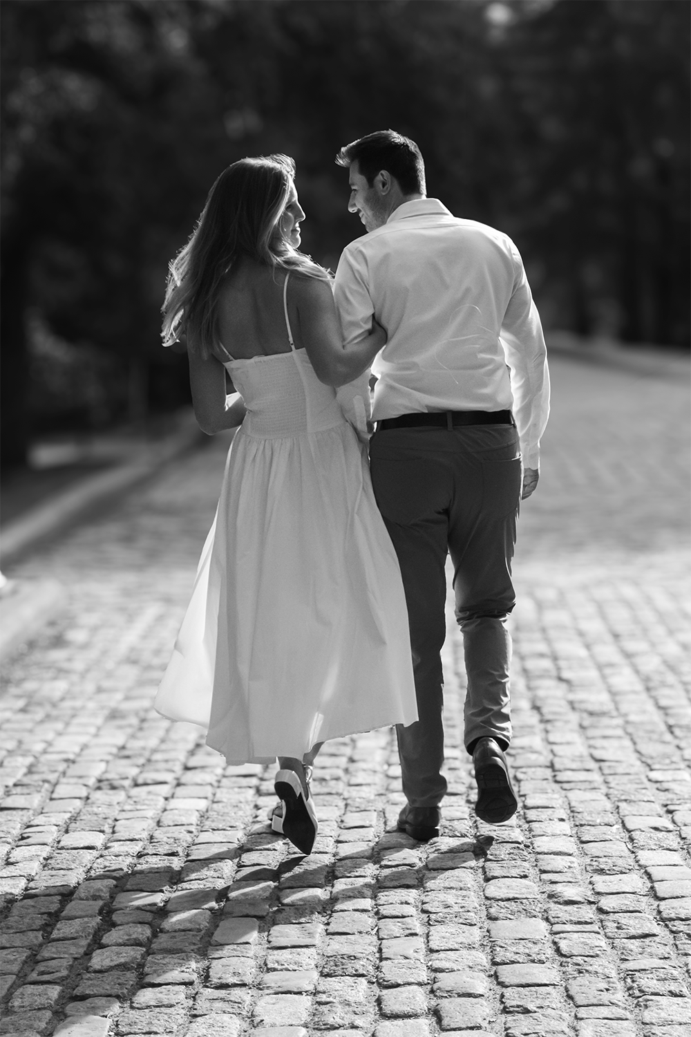 A black and white photo of a couple walking hand in hand on a cobblestone street, smiling and looking at each other.