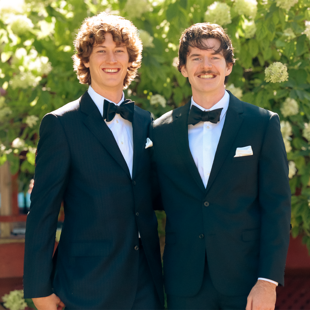 Two men in black tuxedos with bow ties smiling outdoors against green foliage.