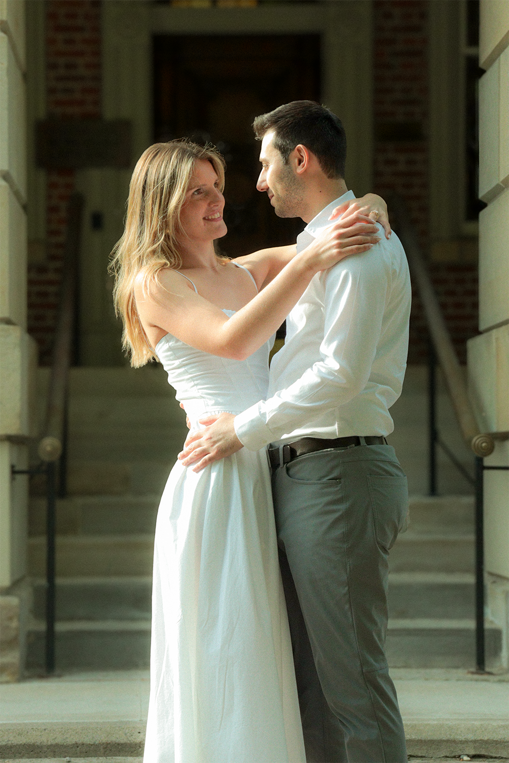 A couple dancing closely on steps outside a building, smiling at each other, with the woman wearing a white dress and the man in a white shirt and dark trousers.