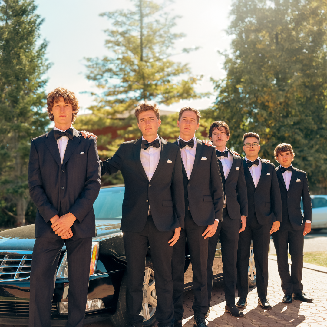 Group of six young men in tuxedos standing outdoors in front of a black luxury car with trees in the background.