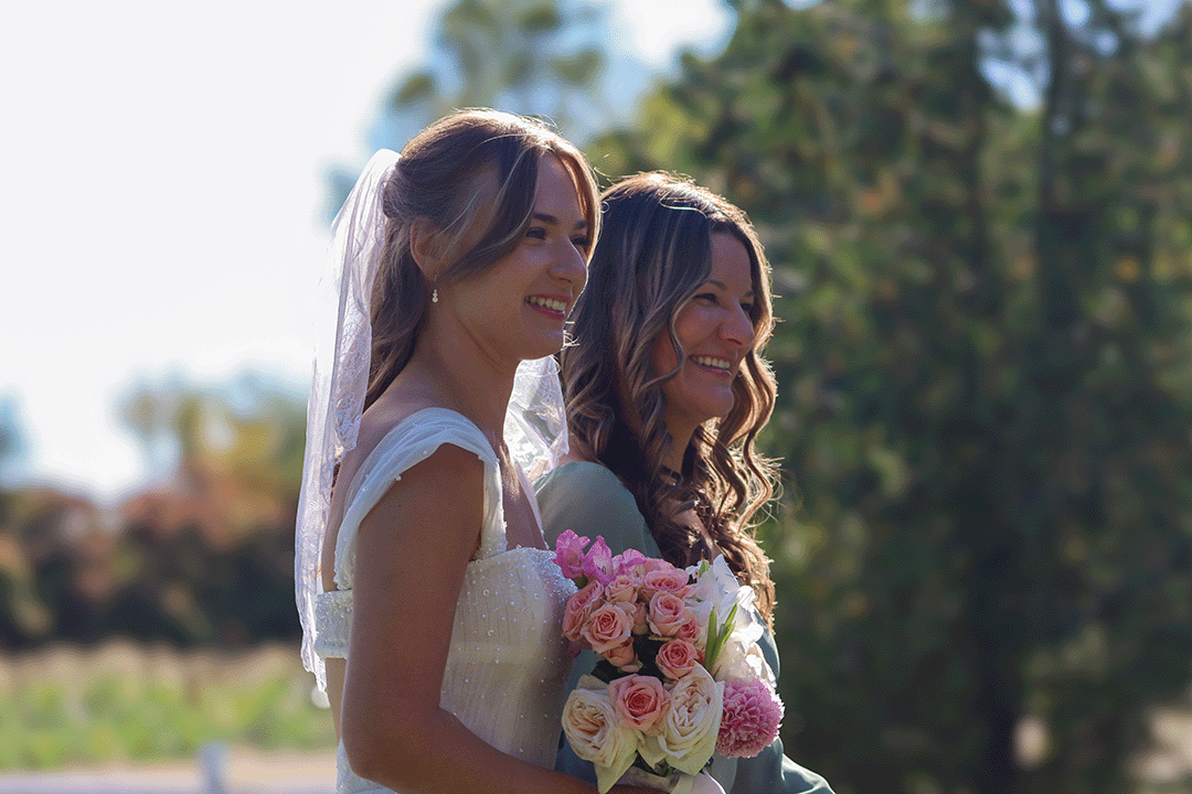 Two women, one wearing a white wedding dress with a veil and holding a bouquet of pink and white flowers, and the other wearing a light-colored dress, are smiling outdoors with trees and a clear sky in the background.
