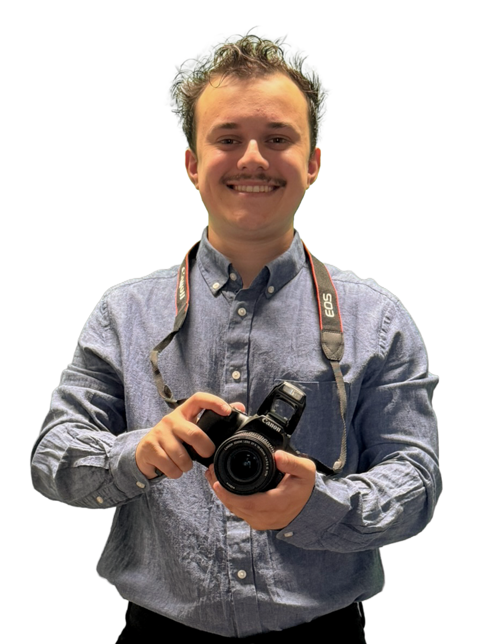 Young man with curly hair, smiling, holding a black Canon camera, wearing a gray button-down shirt with a camera strap around his neck.