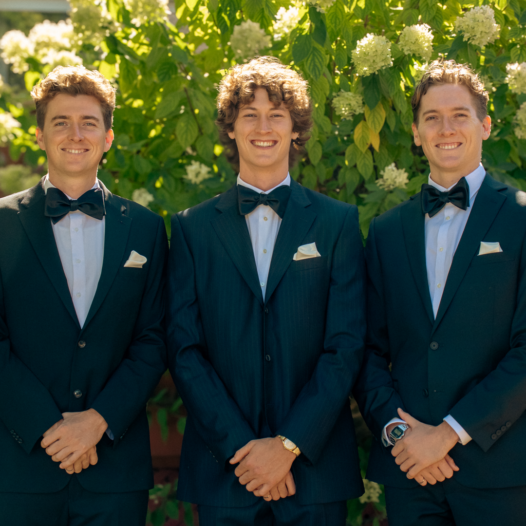 Three young men dressed in tuxedos with bow ties and pocket squares, standing outdoors in front of lush green foliage and white flowers.