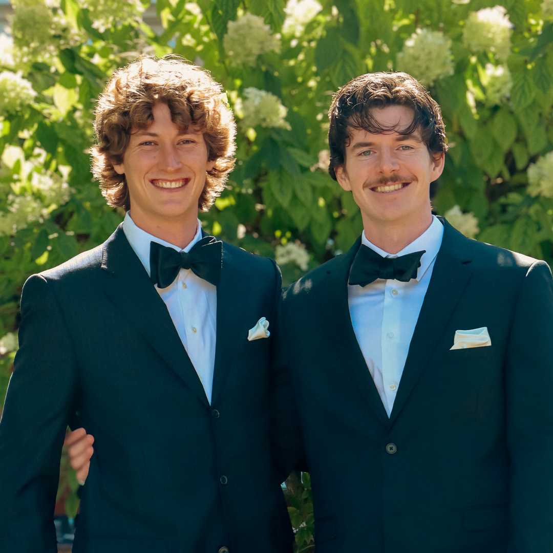 Two men wearing black tuxedos with bow ties and white shirts, smiling, standing outdoors in front of green foliage and white flowers.