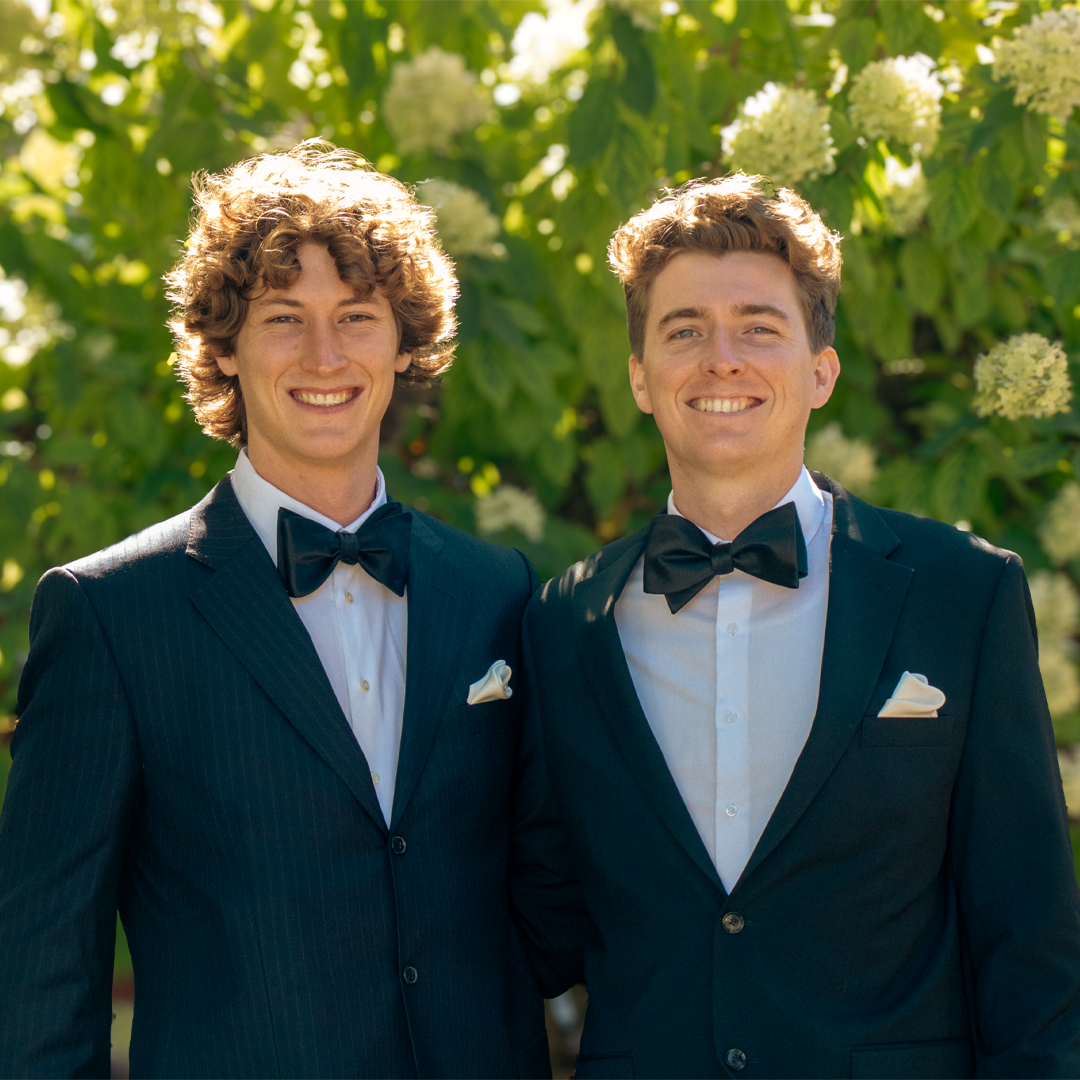 Two young men in tuxedos with bow ties, smiling outdoors with green leafy background.