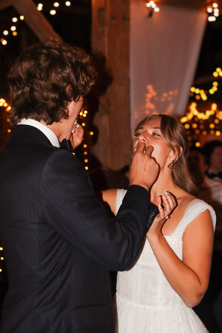 A groom in a black tuxedo feeds cake to a bride in a white wedding dress during their wedding reception.