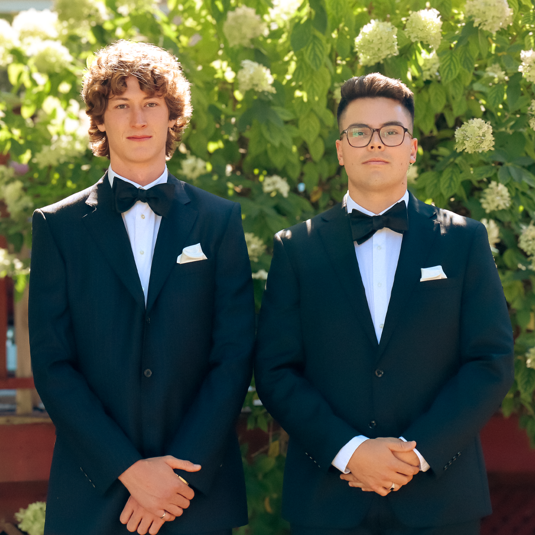 Two young men in black tuxedos with white shirts and black bow ties standing outdoors in front of green foliage and white flowers.