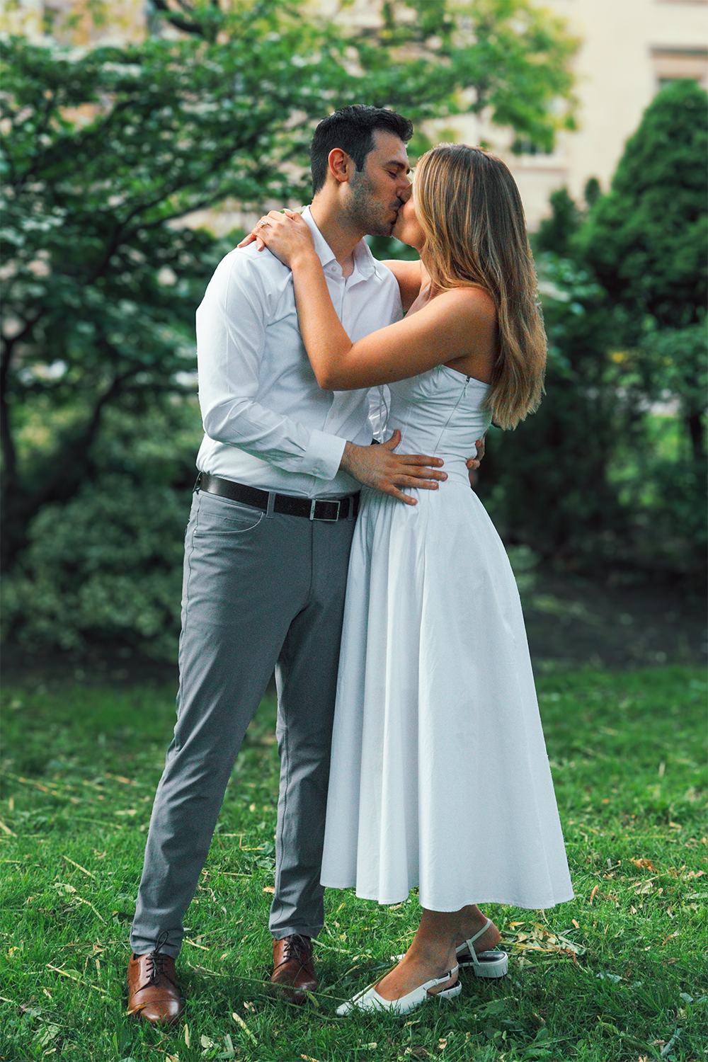 A couple dressed in white, sharing a kiss in a park with green trees in the background.