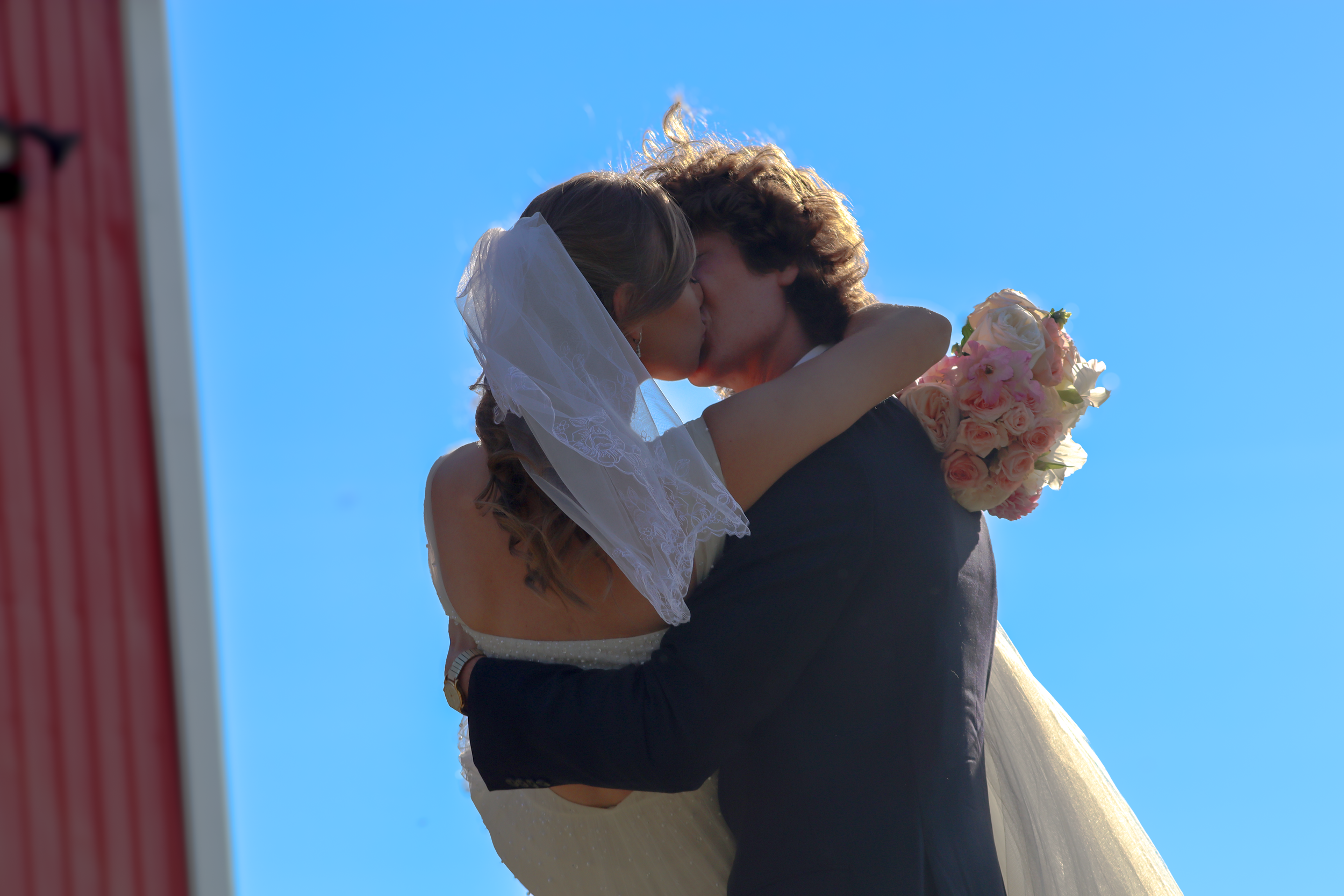 A bride and groom are sharing a kiss outdoors against a clear blue sky. The bride is wearing a white wedding dress and veil, holding a bouquet of pink and white roses. The groom is dressed in a dark suit.