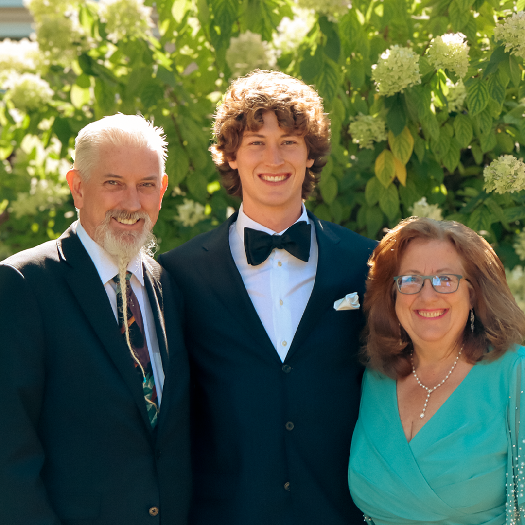 A young man in a tuxedo with a bow tie standing between a man with a white beard and a woman with glasses, all smiling outdoors in front of green plants.