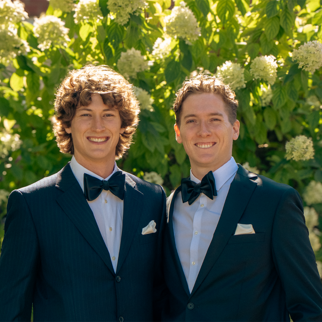 Two men wearing tuxedos with bow ties, standing outdoors in front of a lush green bush with white flowers, smiling at the camera.