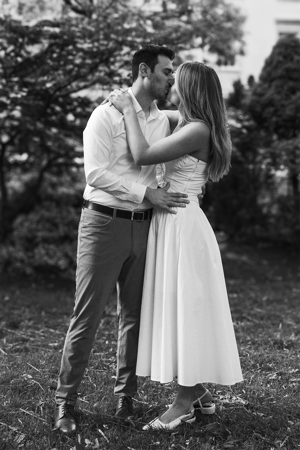 A black and white photo of a couple kissing outdoors. The man is wearing a dress shirt and pants, and the woman is in a sleeveless dress. The woman's arms are around the man's neck, and the man's hand is on the woman's waist. They are standing on a g