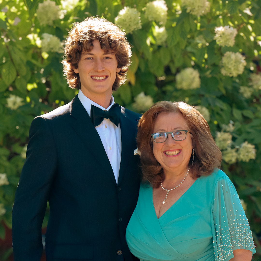 A young man in a tuxedo with a bow tie standing next to an older woman in a turquoise dress with pearl jewelry, both smiling outdoors in front of green foliage and white flowers.
