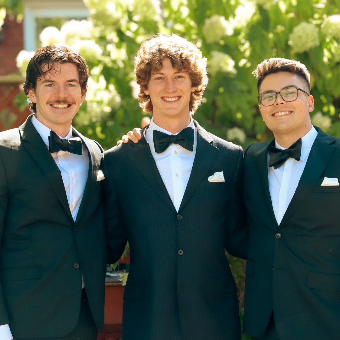 Three young men in black tuxedos and bow ties smiling outdoors, with green foliage in the background.