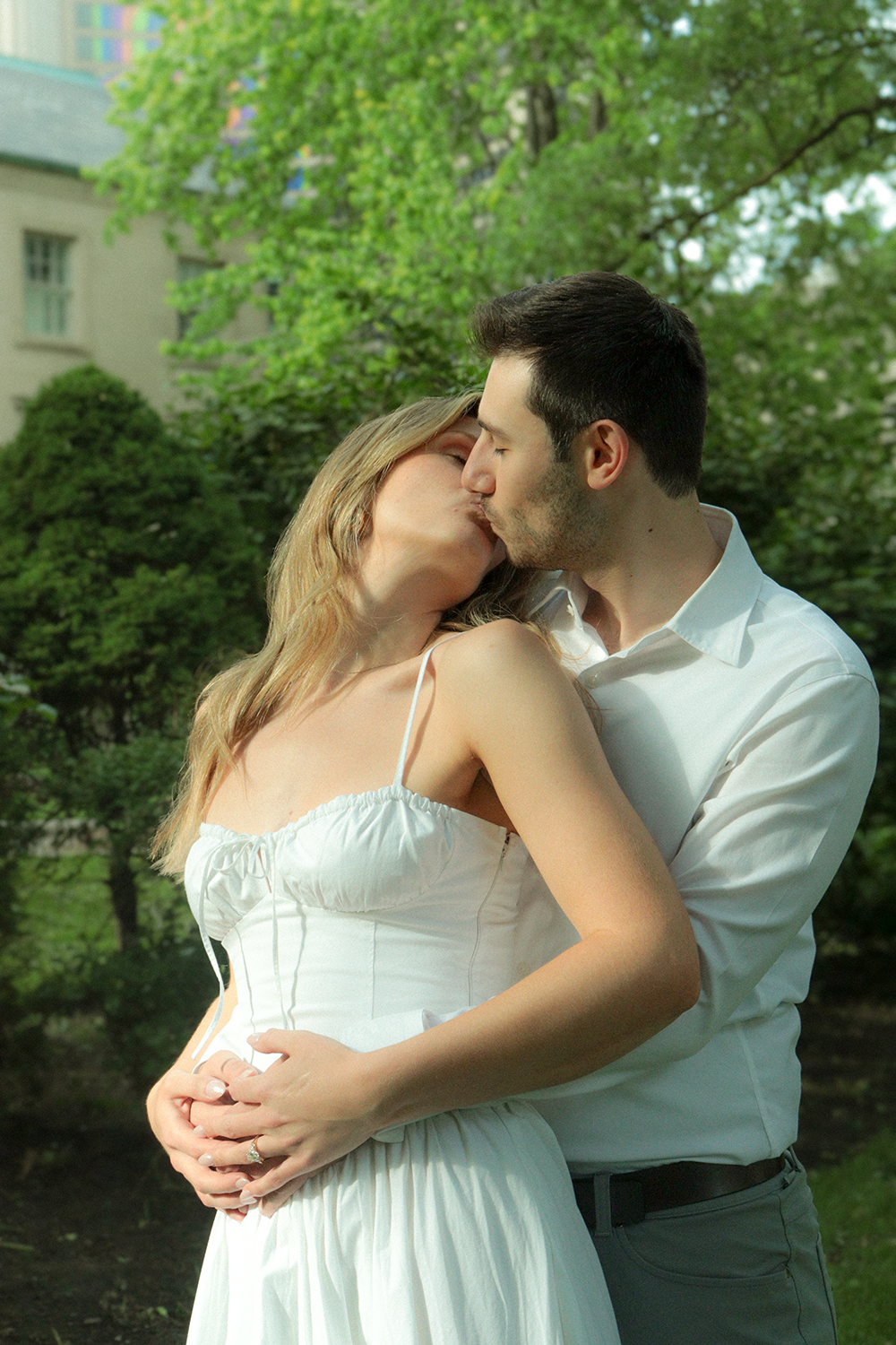 A couple sharing a kiss outdoors surrounded by green trees.