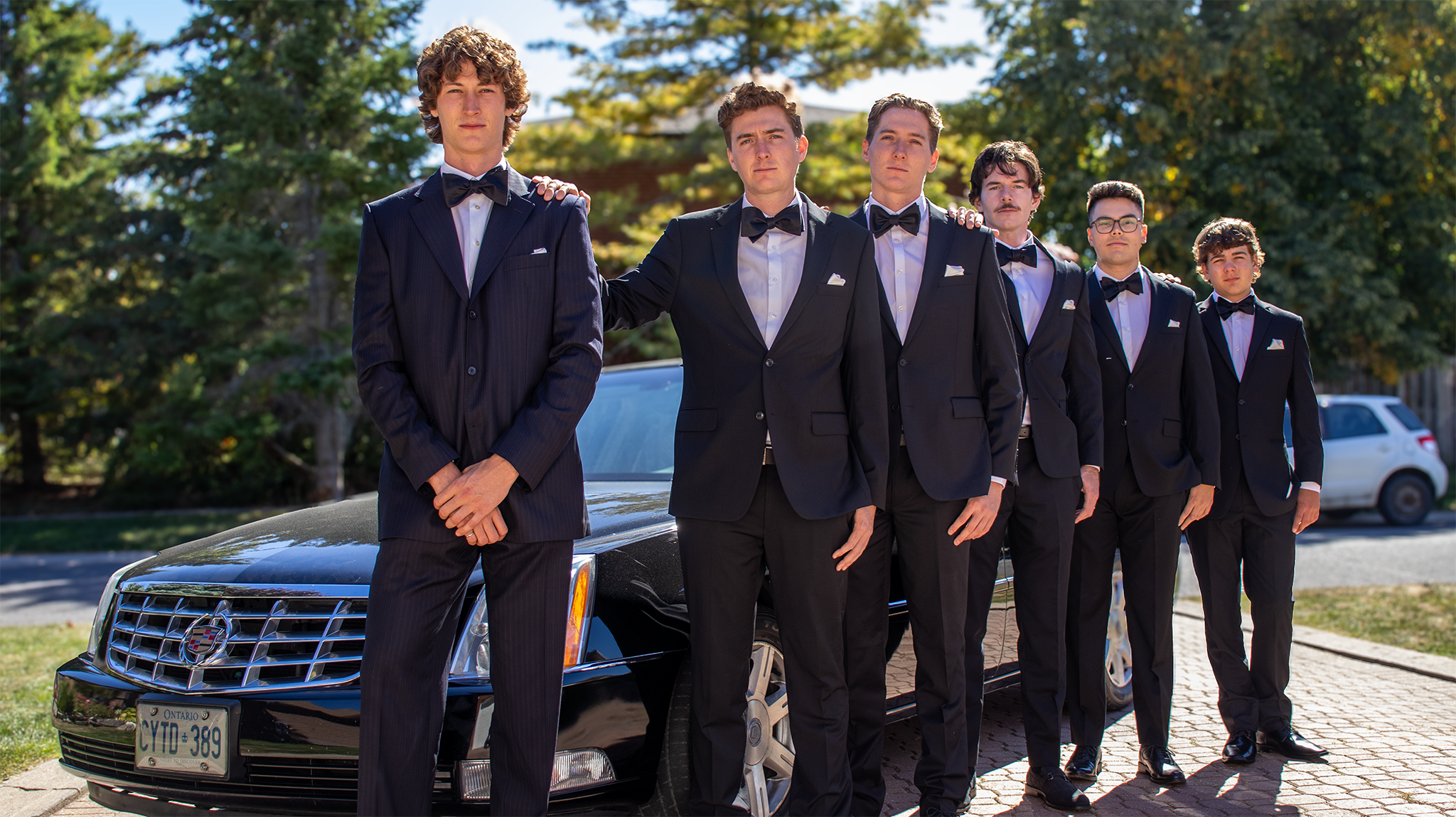 Six young men dressed in black tuxedos with bow ties, standing in a line in front of a black luxury car outdoors, with trees and cars in the background.