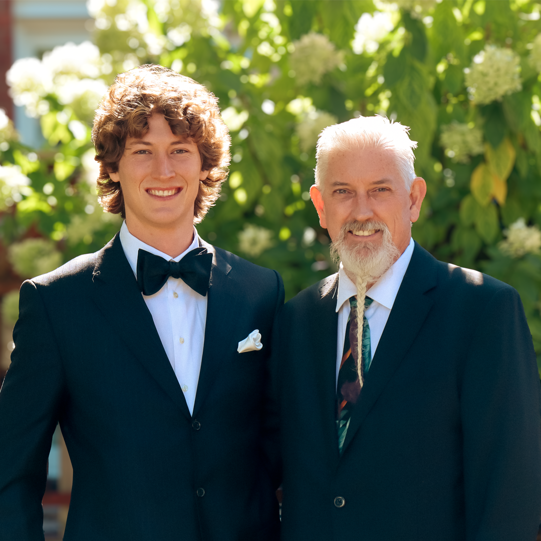 Two men dressed in suits, one young with curly hair and a bow tie, the other older with a beard and tie, standing outdoors with green foliage and white flowers in the background.