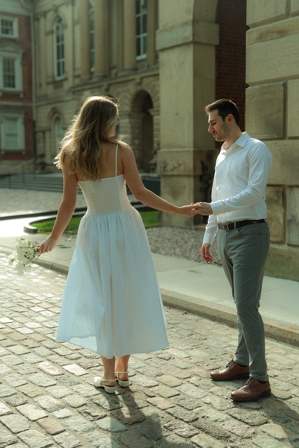 A man and woman holding hands on a cobblestone street, woman in a white dress holding flowers, man in a white shirt and gray pants, outdoor urban setting with historic building.