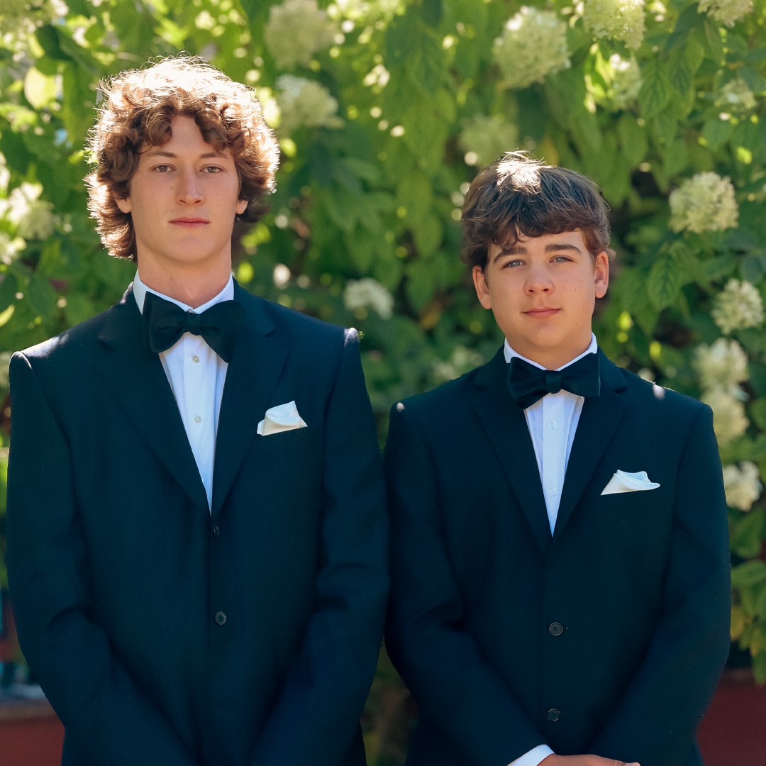 Two young men in tuxedos with bow ties and white pocket squares standing outdoors in front of green leafy bushes with white flowers.