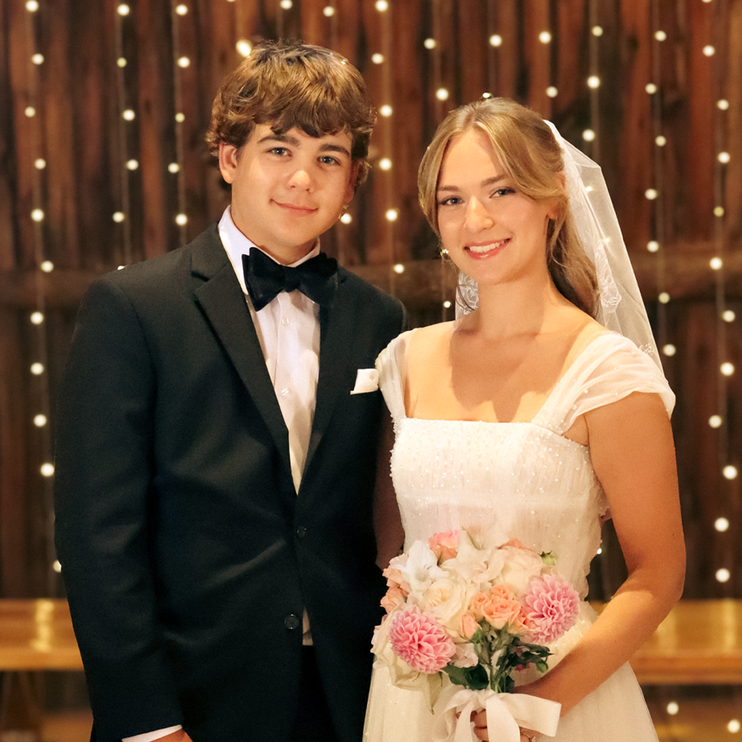 A young man and woman dressed in wedding attire, smiling for a photo at a wedding reception or ceremony.