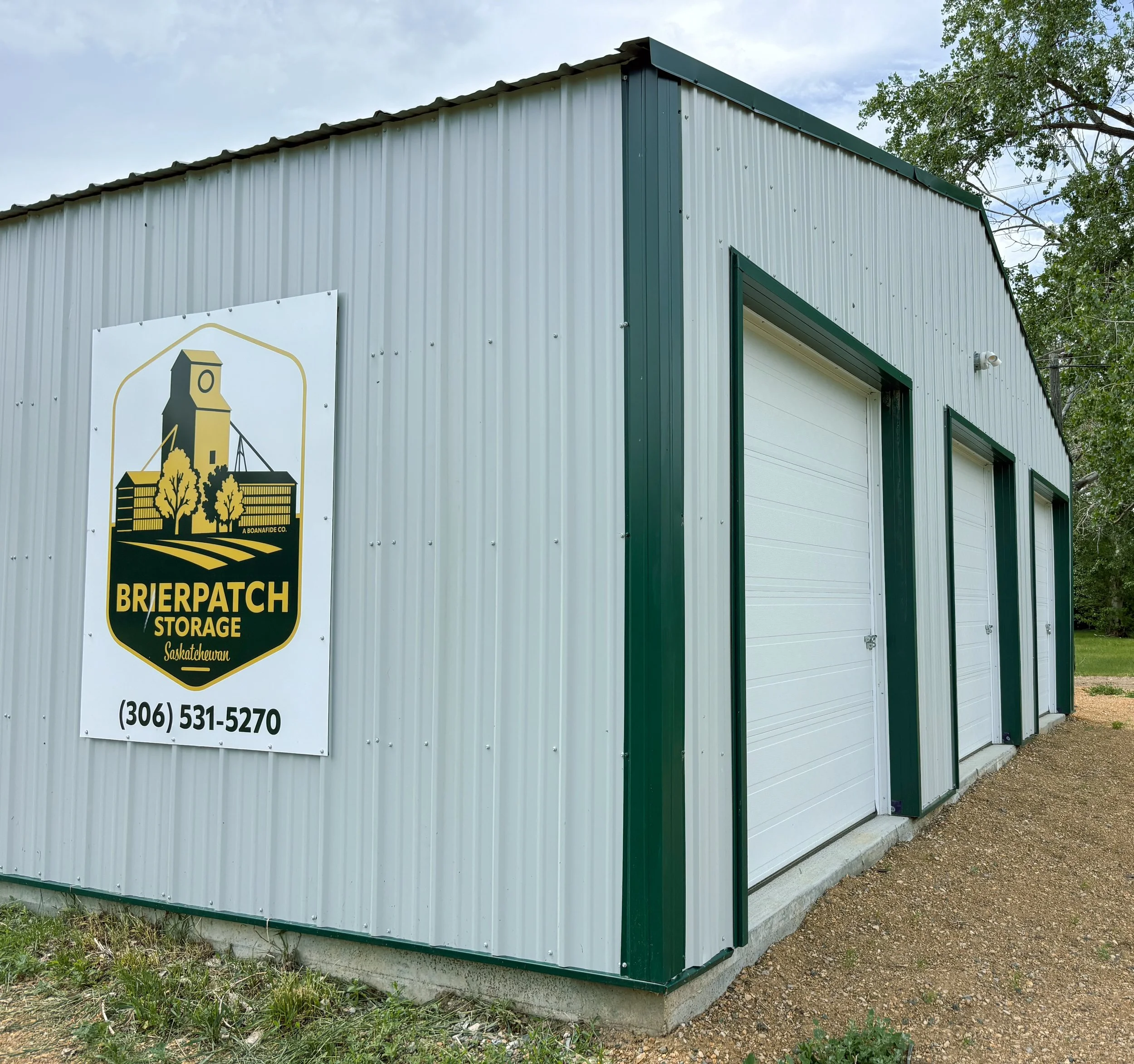 Metal storage building with a white exterior and green trim, featuring a sign for Brierpatch Storage in Saskatchewan, with trees and a cloudy sky in the background.