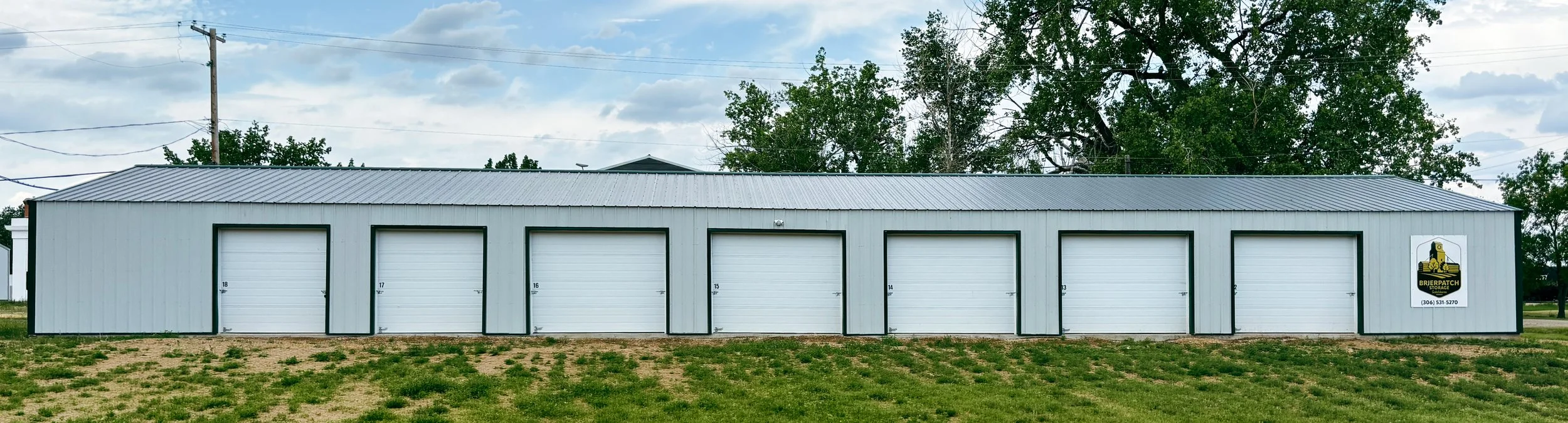 A row of six gray storage units with white doors, numbered 18 to 13 from left to right, situated on a grassy area with a cloudy sky and trees in the background. There is a sign on the far right featuring a yellow and black logo and contact information.