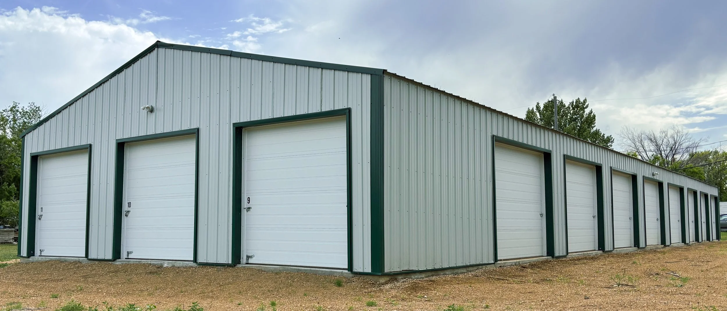 A row of storage units or garages with white doors and green trim, set outdoors on a dirt surface with trees and a cloudy sky in the background.
