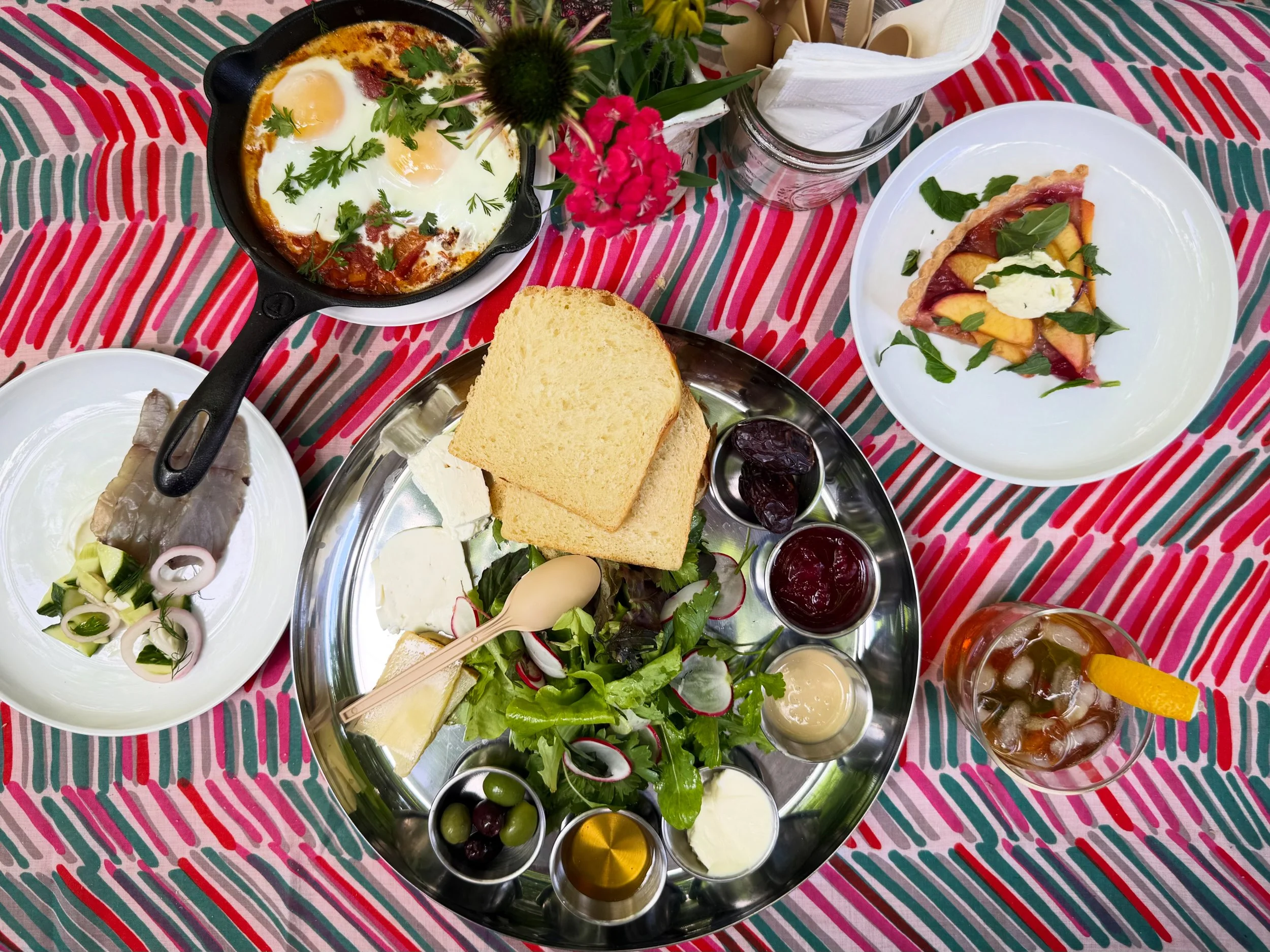 A table set with a variety of breakfast foods including a skillet of eggs and vegetables topped with herbs, slices of bread, fresh salad greens with radishes, small jars of jam or jelly, slices of cheese, and drinks, all on a colorful, striped tablecloth.