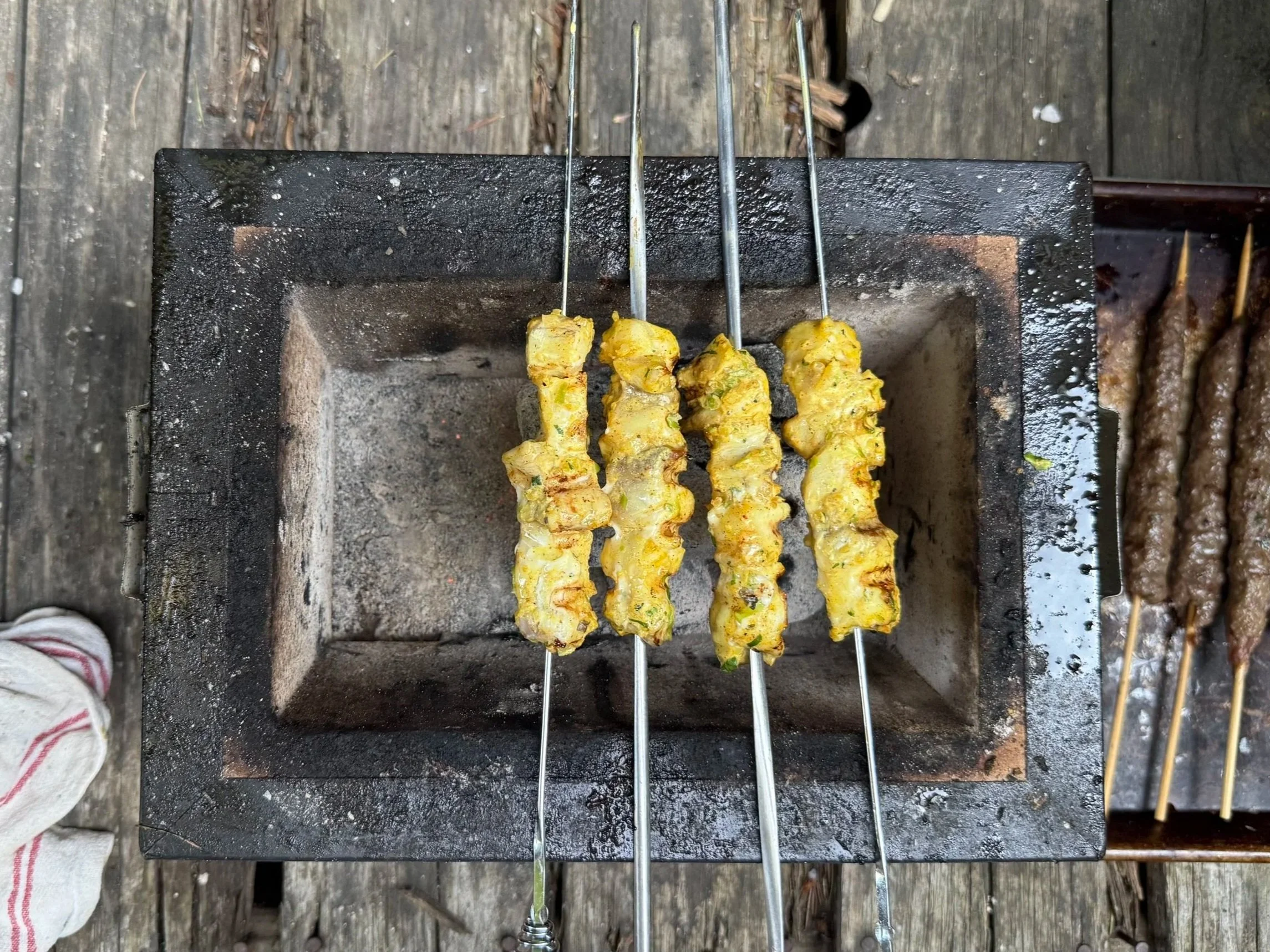 Shish kebabs cooking on metal skewers over a charcoal grill, with some chicken and vegetables visible.