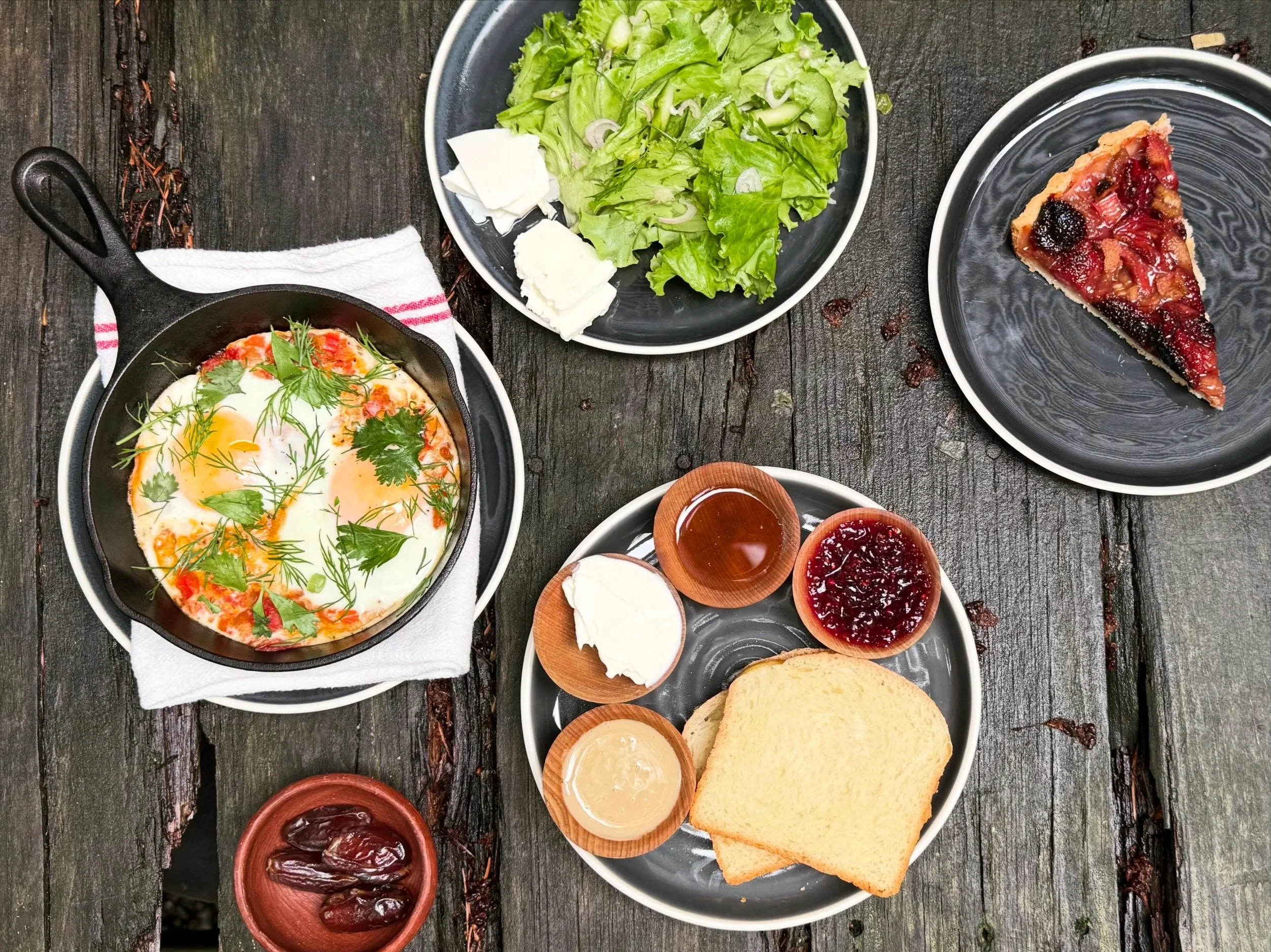 Breakfast spread on a rustic wooden table including a skillet with eggs garnished with herbs, a salad with lettuce, cucumbers, and onions, slices of bread, and small bowls of jam and syrup.