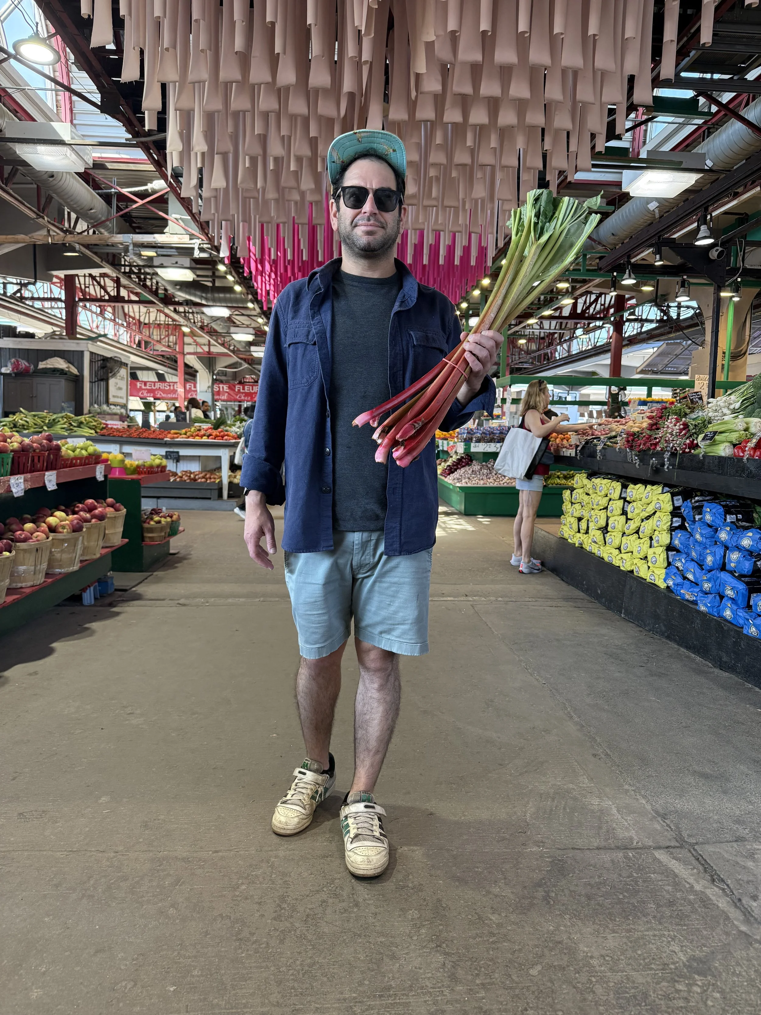 A man in casual clothes and sunglasses holding bunches of rhubarb in a grocery store.