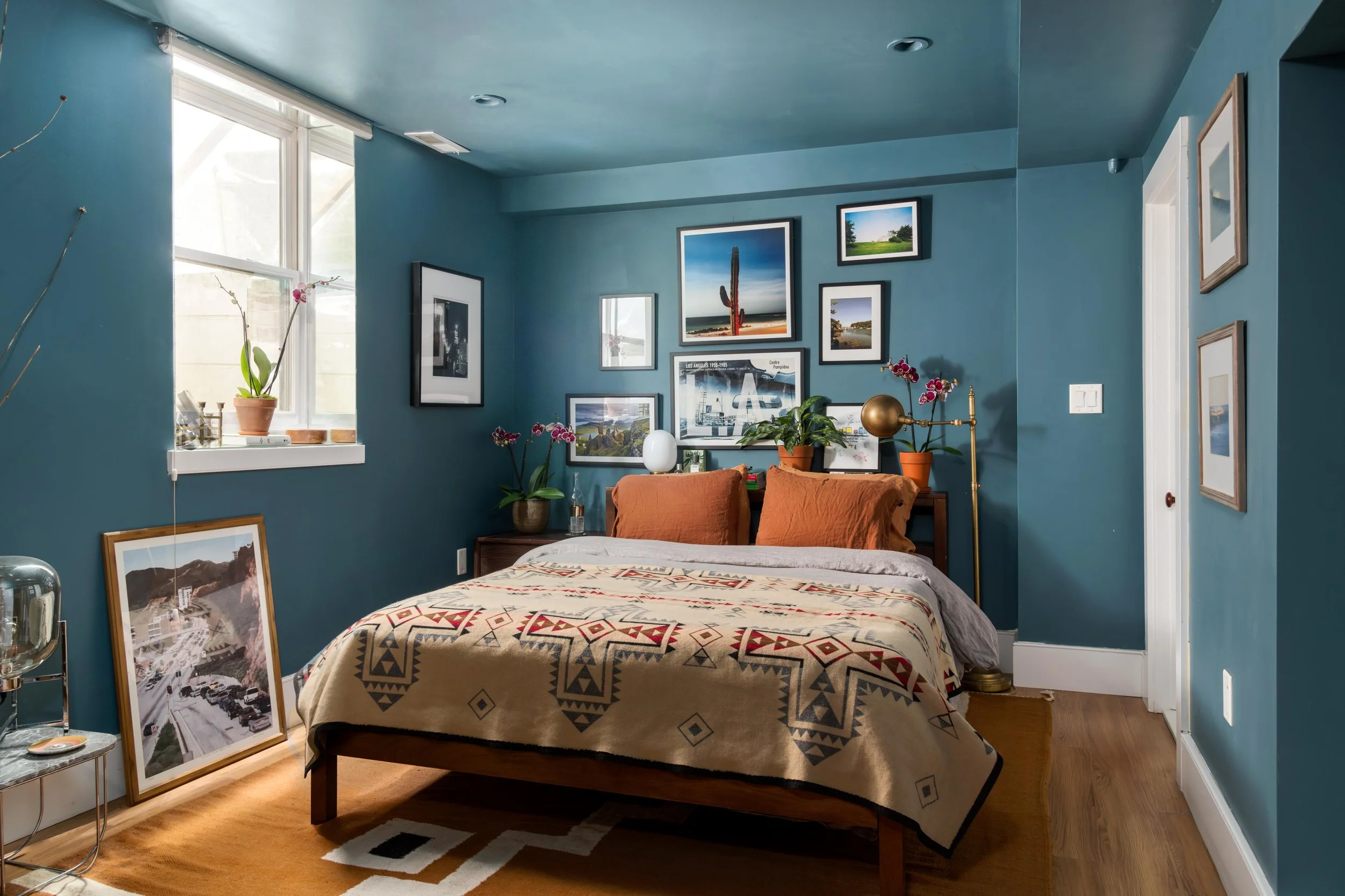 A cozy bedroom with teal walls decorated with framed artwork and windows providing natural light. The bed has a patterned blanket and orange pillows. Various potted plants and floor art rest against the wall.