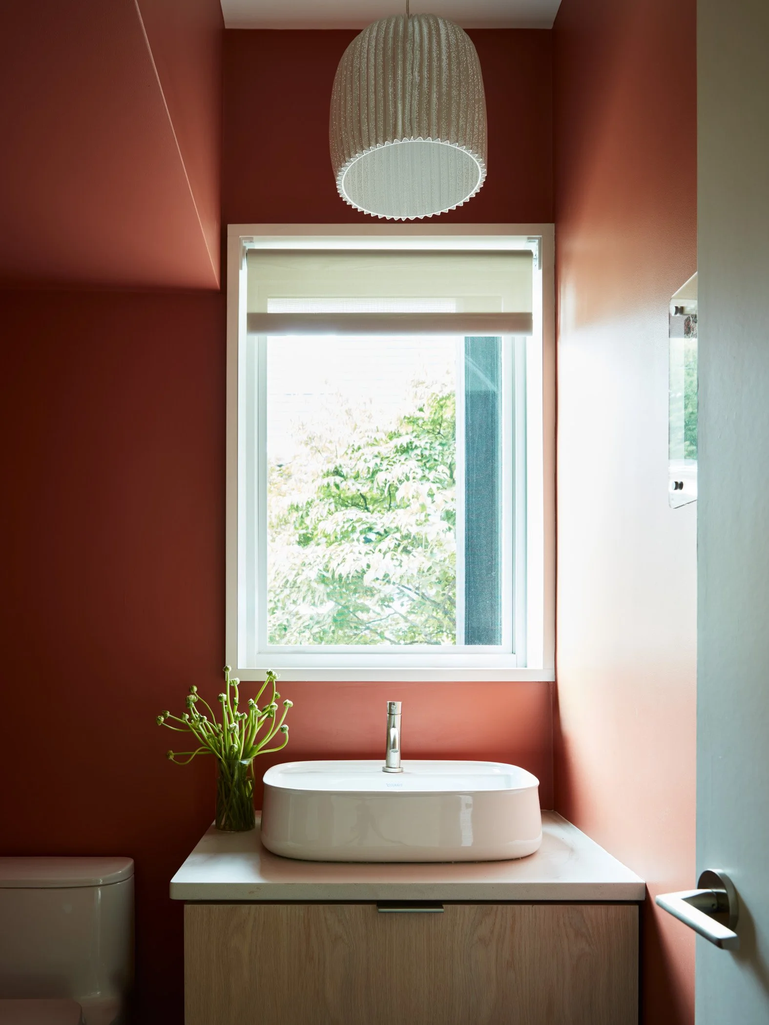 Bathroom with red walls, a large window with a beige blind, a white vessel sink on a light wood vanity, a green plant in a glass vase to the side, and a white toilet to the left. A ceiling-mounted light fixture is above.