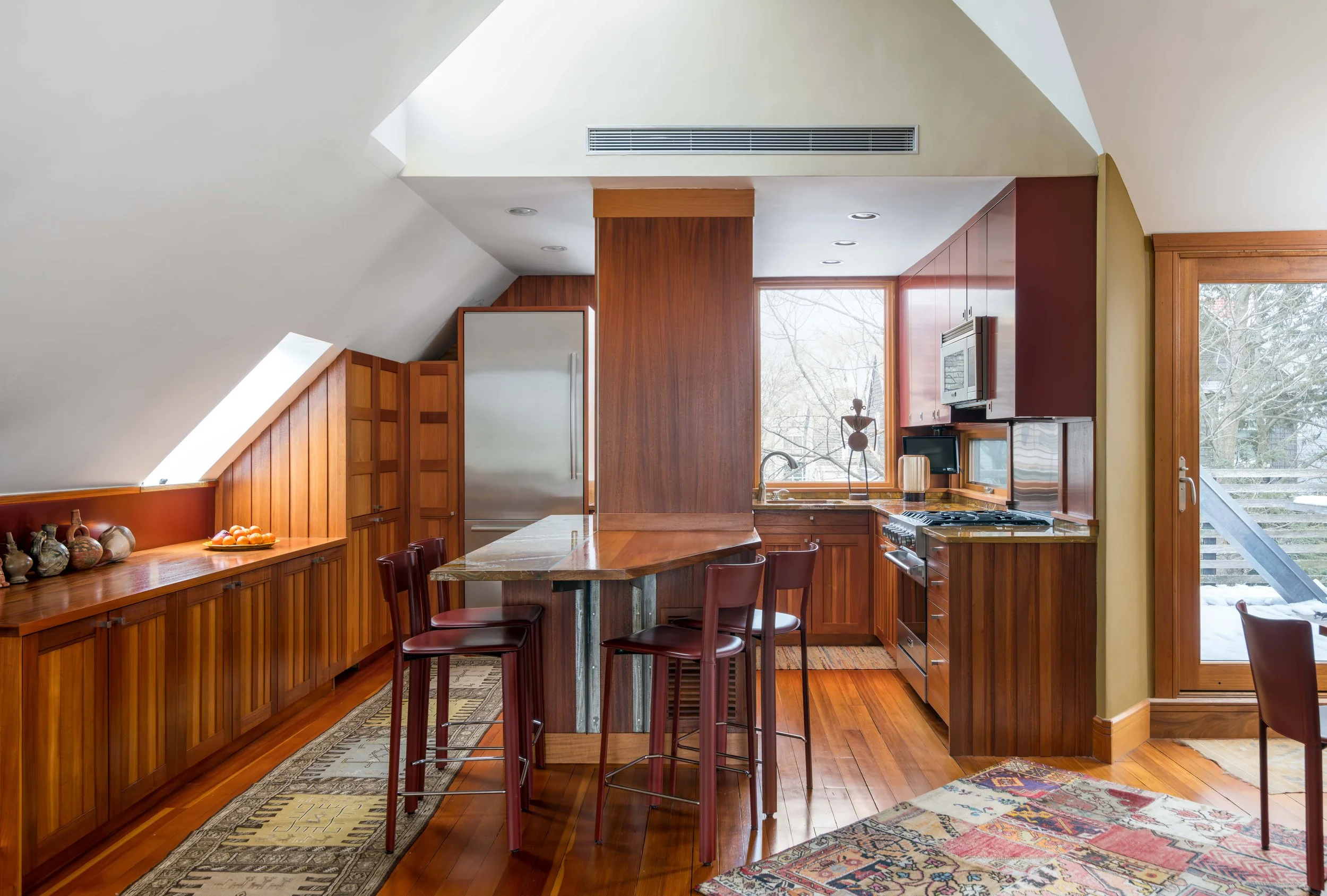 A kitchen with wooden cabinets, a marble-topped island, a large window with trees outside, and a skylight bringing in natural light.