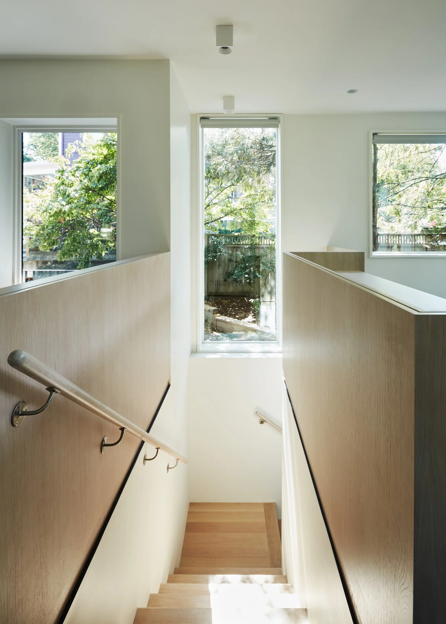 Indoor staircase with wooden steps, beige walls, and handrails on both sides leading down to a landing with light-colored wooden flooring, illuminated by natural light from large windows and a glass door at the top of the stairs.