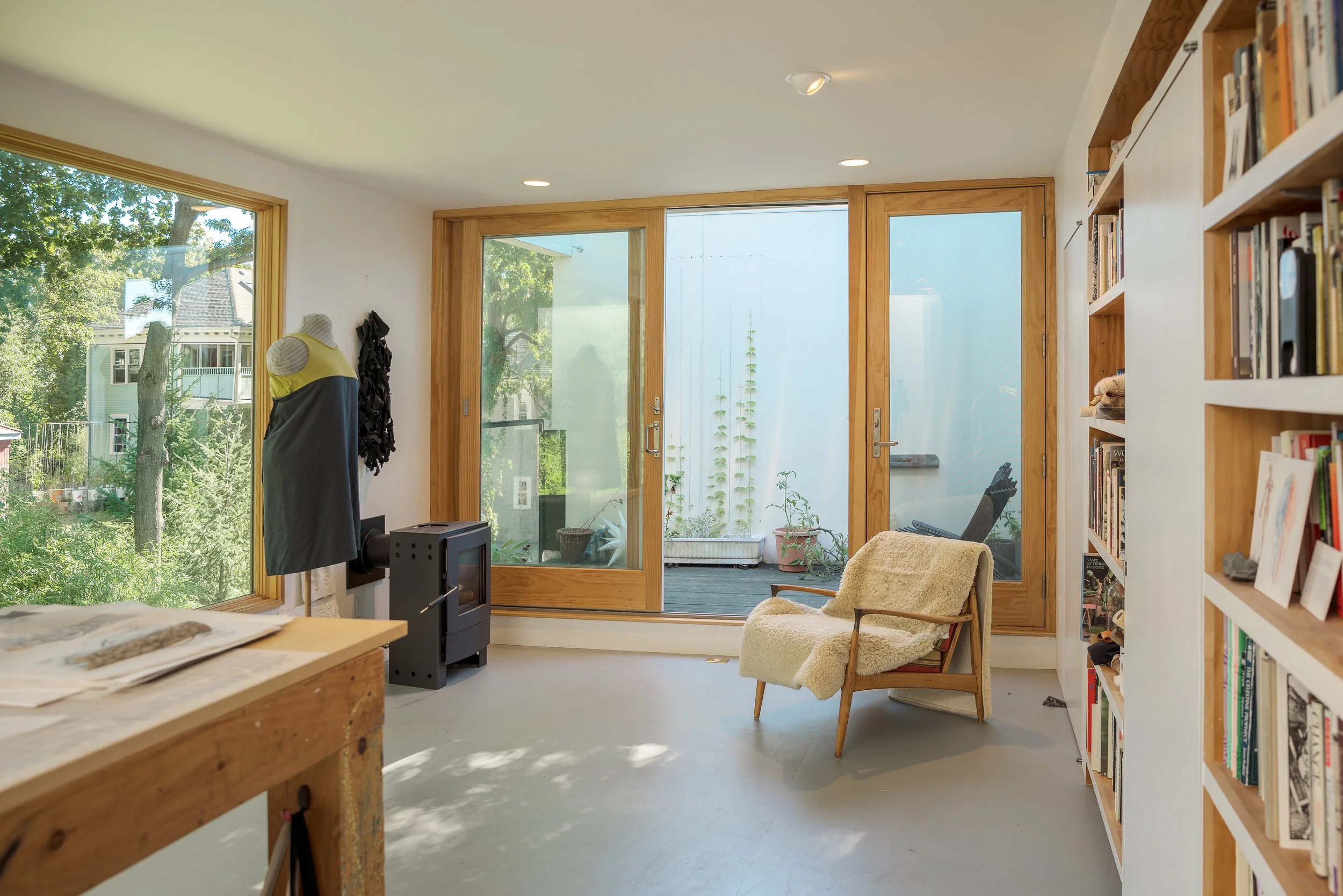 A cozy interior room with large wooden-framed glass sliding doors leading to an outdoor deck with potted plants. Inside, a cream-colored armchair with a wooden frame is placed near a white bookshelf filled with books. The room has white walls and cei