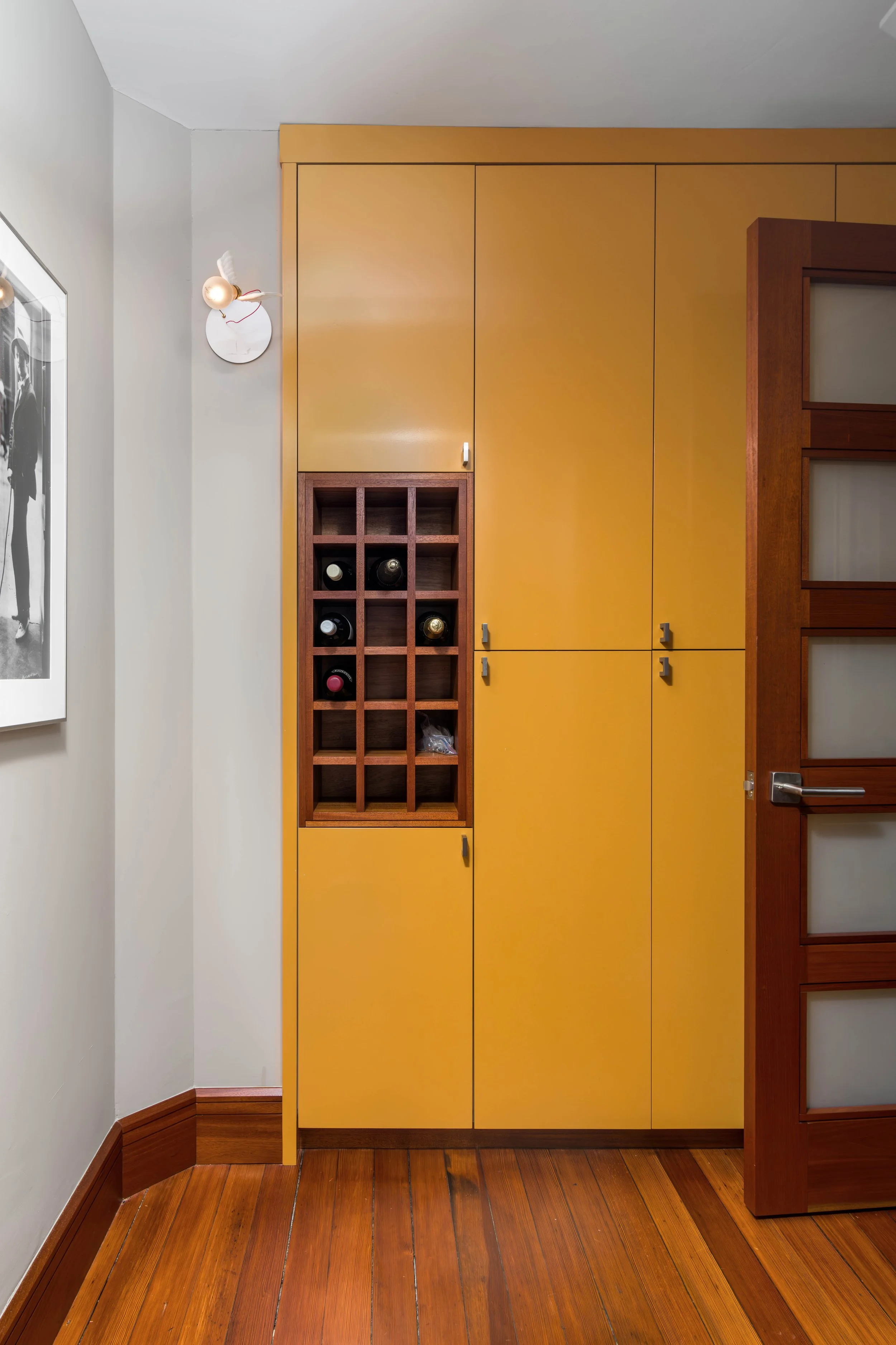A corner of a room with white walls, a wooden floor, and yellow cabinets. There is a wooden wine rack with bottles on the wall and a circular wall clock above it. A framed black-and-white photograph is partially visible on the left side.