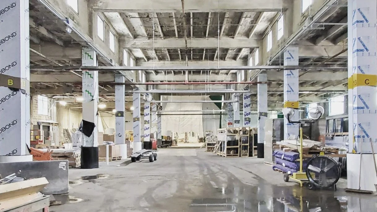 Construction site of a large building interior with exposed ceiling beams, columns, and scattered construction materials and tools.