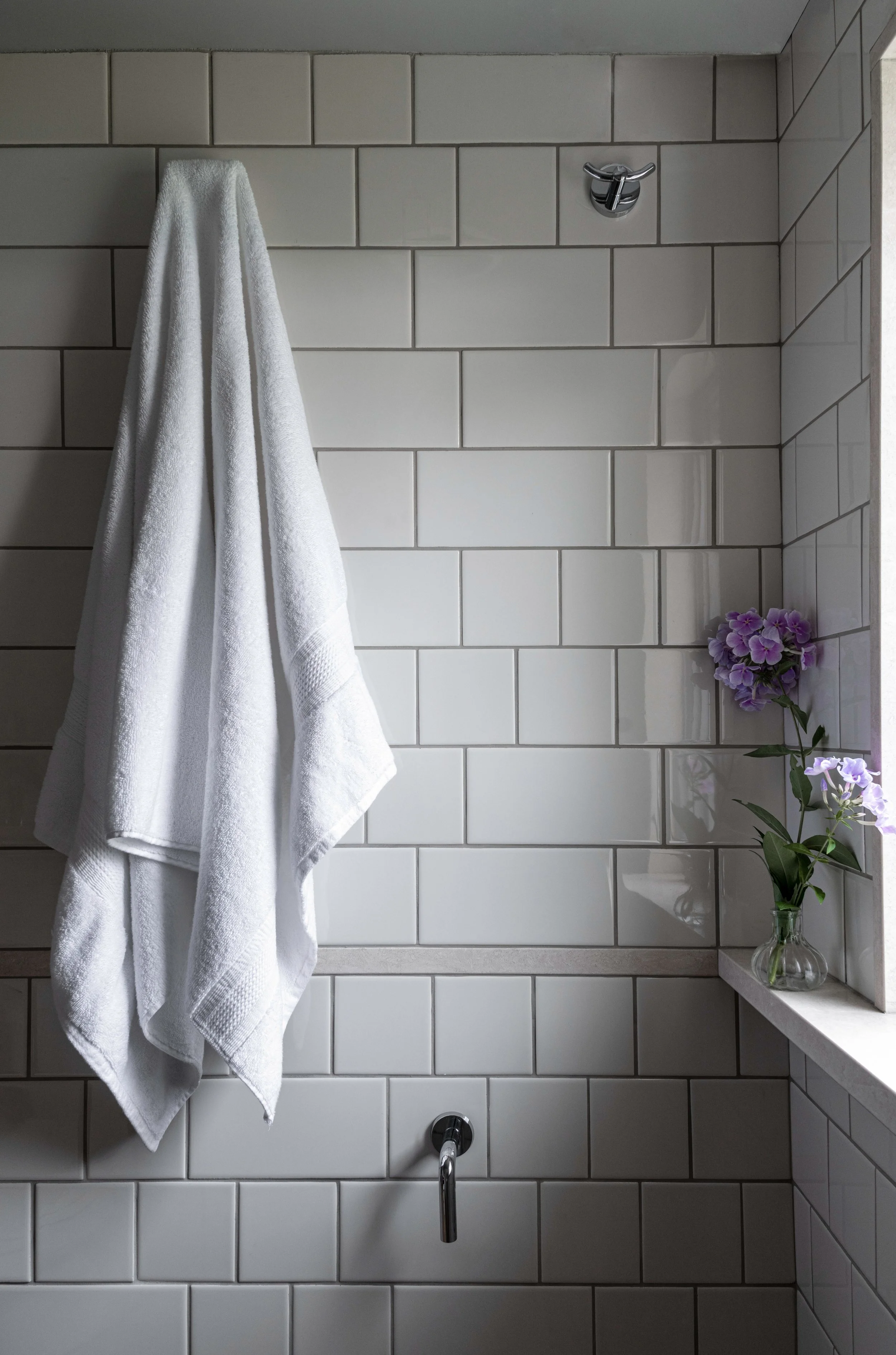White towel hanging on a hook on a tiled bathroom wall, with purple flowers in a vase on a windowsill.