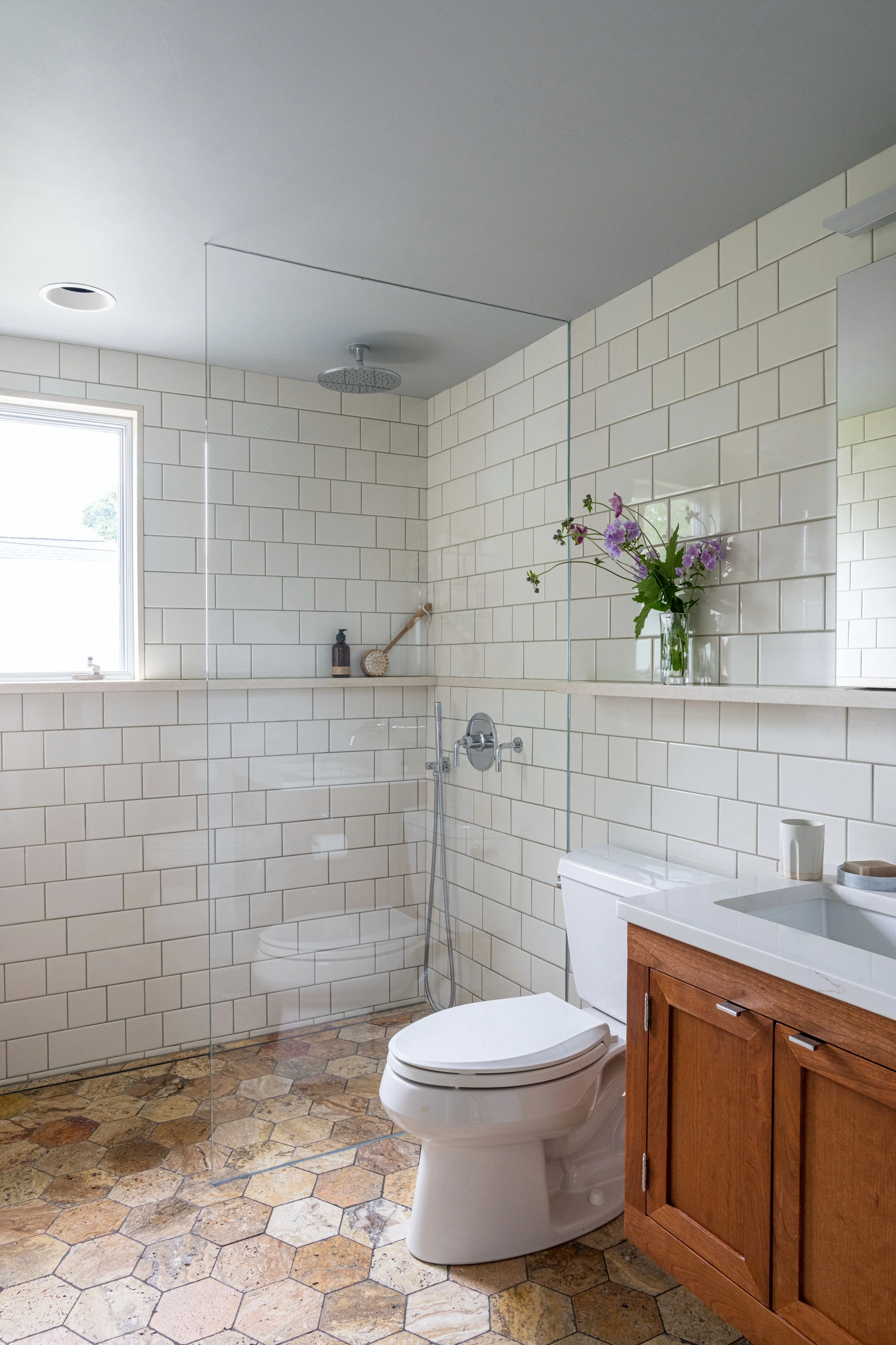 Modern bathroom with tiled walls, a floating glass shower enclosure, a window, a toilet, a wooden vanity with a white sink, and a vase of purple flowers on the counter.