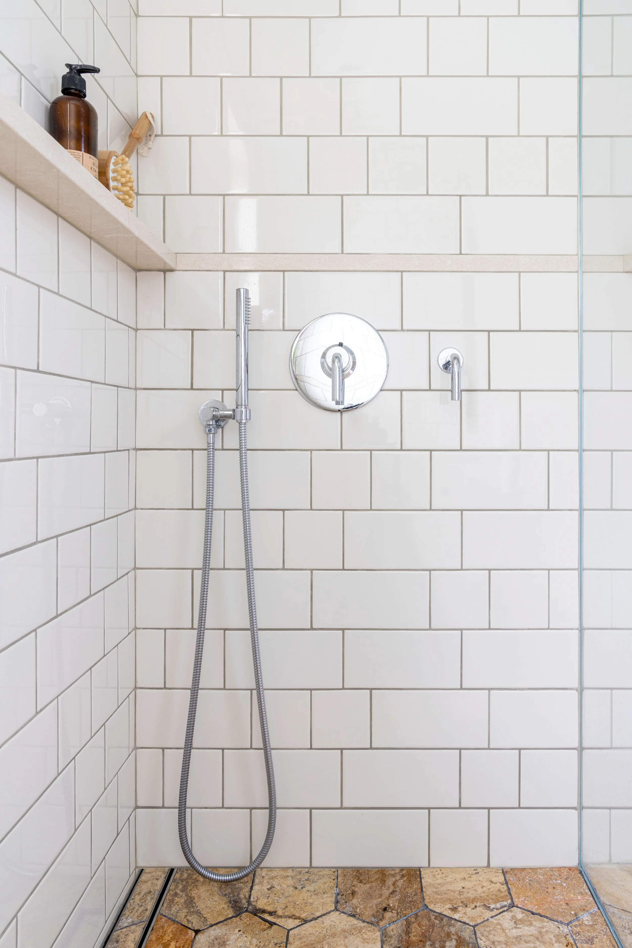 Shower area with white tiled walls, a chrome showerhead with a hose, a round chrome fixture, and a wooden shelf holding a brown soap dispenser, a beige bath brush, and a small white object. The floor has hexagonal beige and brown tiles.