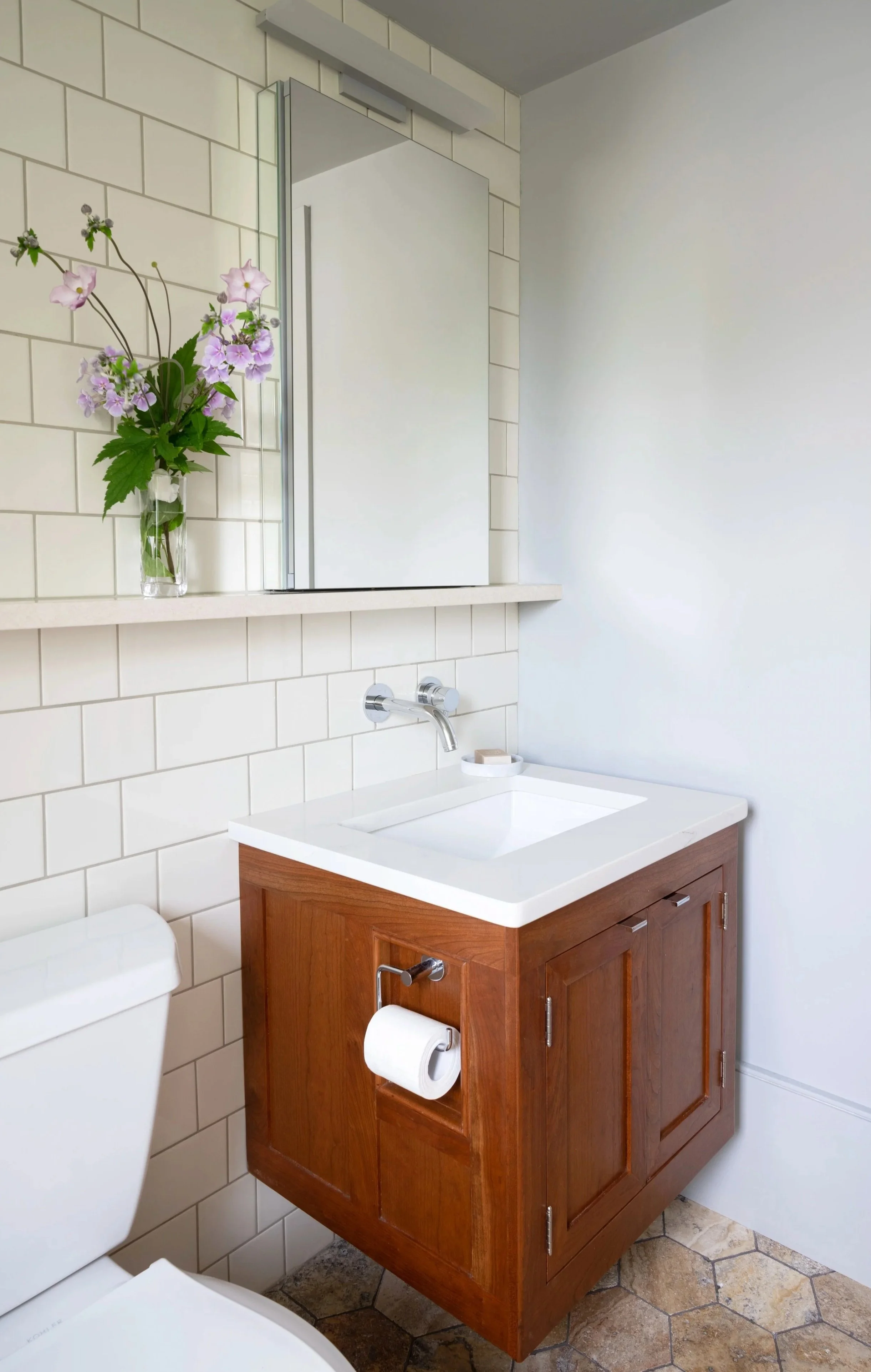 Bathroom with a wooden vanity, white sink, wall-mounted mirror, purple flower arrangement in a glass vase, and a toilet with a toilet paper roll. The wall is tiled with white subway tiles on the left and painted white on the right, with hexagonal sto