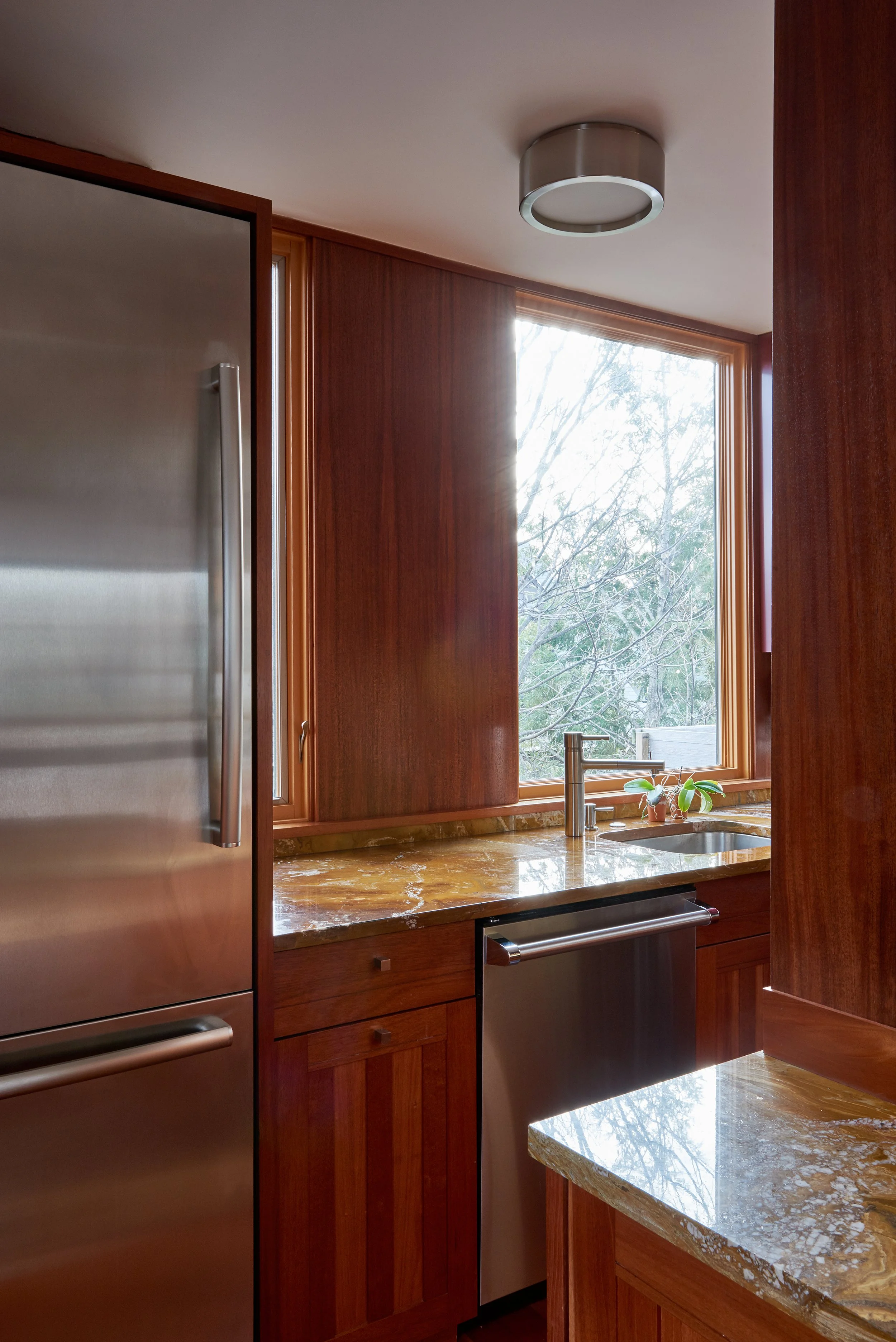 A kitchen featuring a wooden window frame with a large window and a view of trees outside. The kitchen has a polished marble countertop with a small plant near the sink, a modern faucet, and stainless steel appliances including a refrigerator and dis