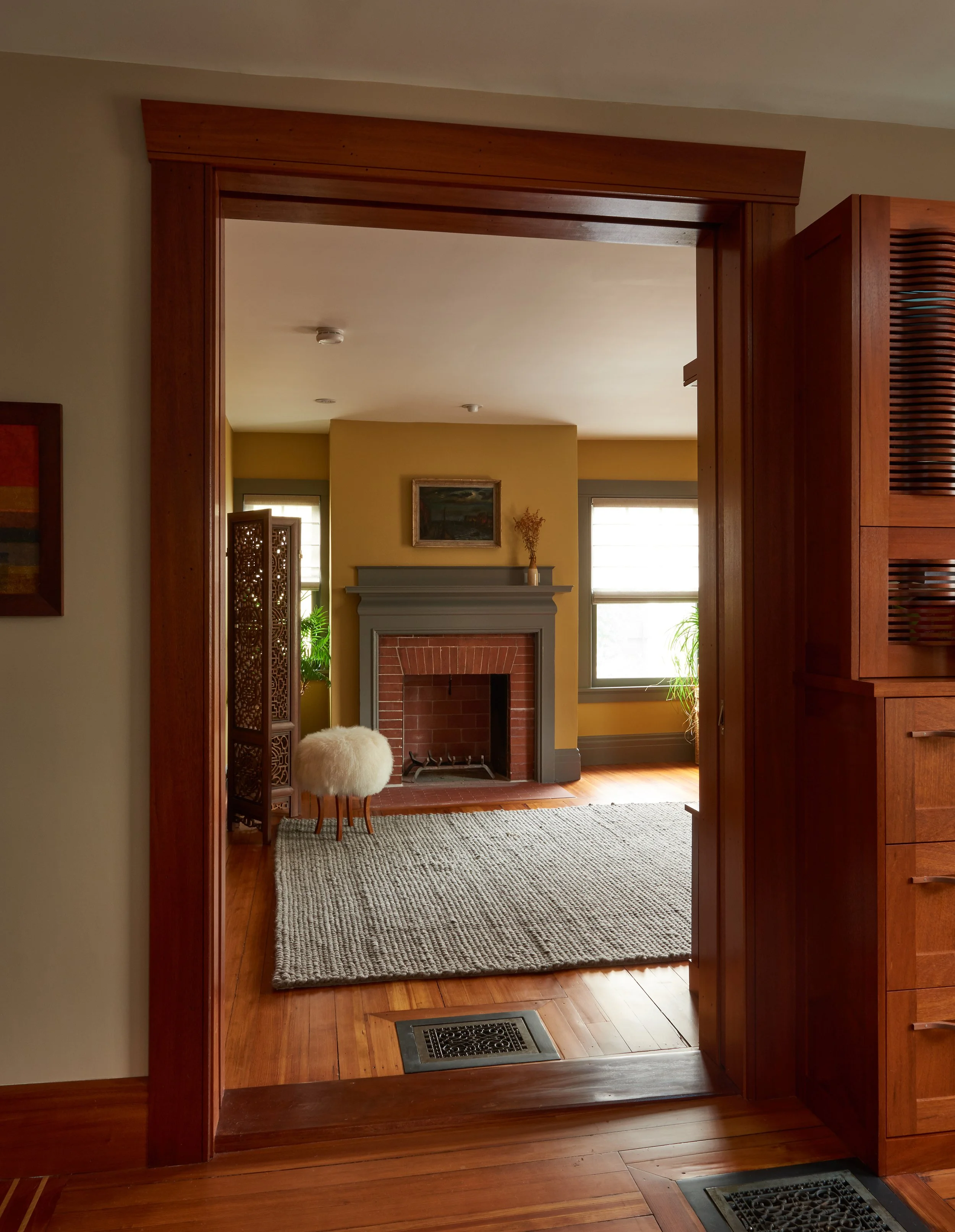View of a living room through a large wooden doorway. The room features a fireplace with a painting above it, two windows with blinds, and various decorative plants. There's a rug on the wooden floor and a fluffy round stool near a wooden room divide