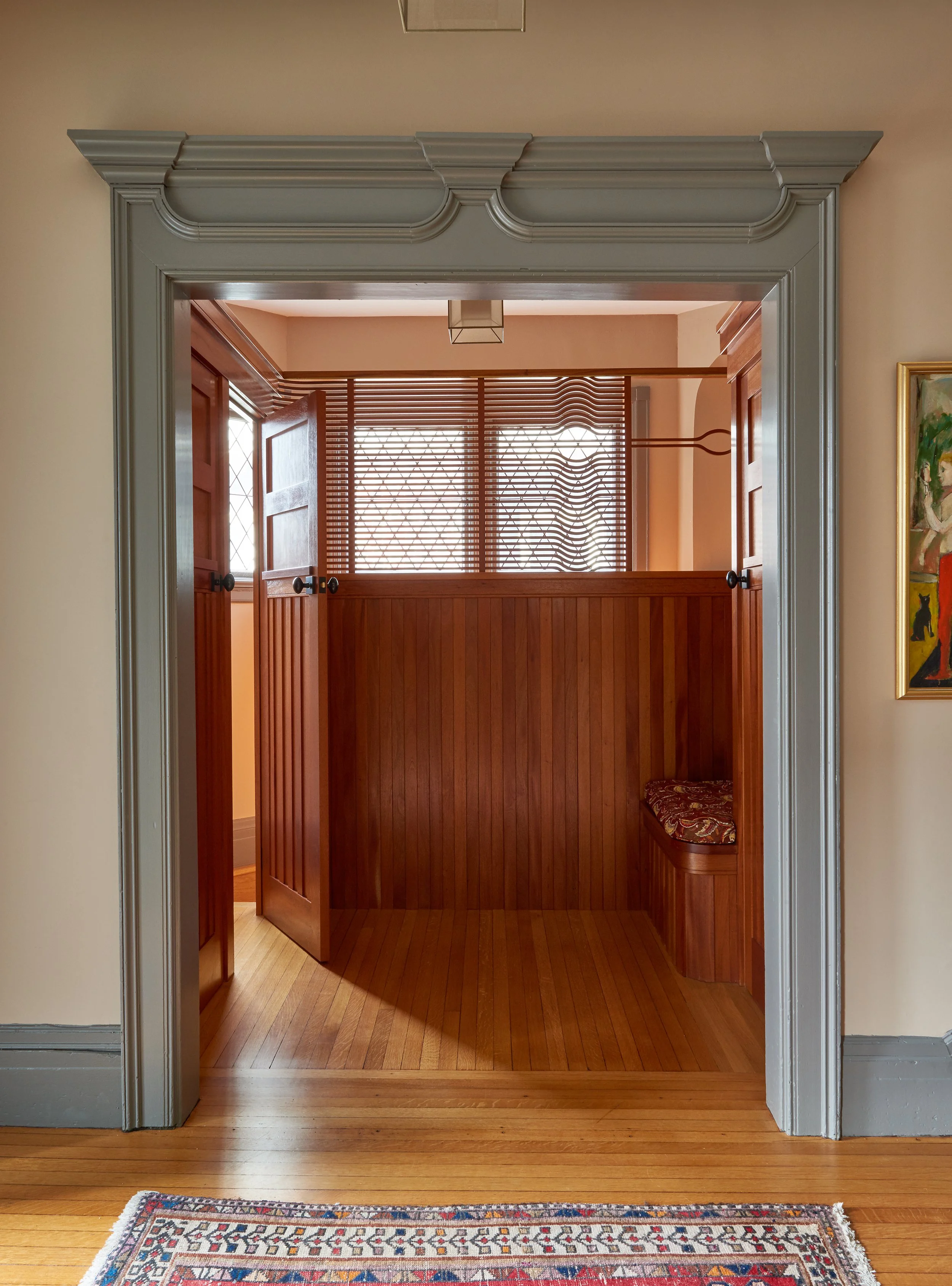 View through a doorway into a room with wooden doors, window with wooden blinds, and a built-in seat with patterned cushion; hardwood floor and a colorful rug at the entrance.