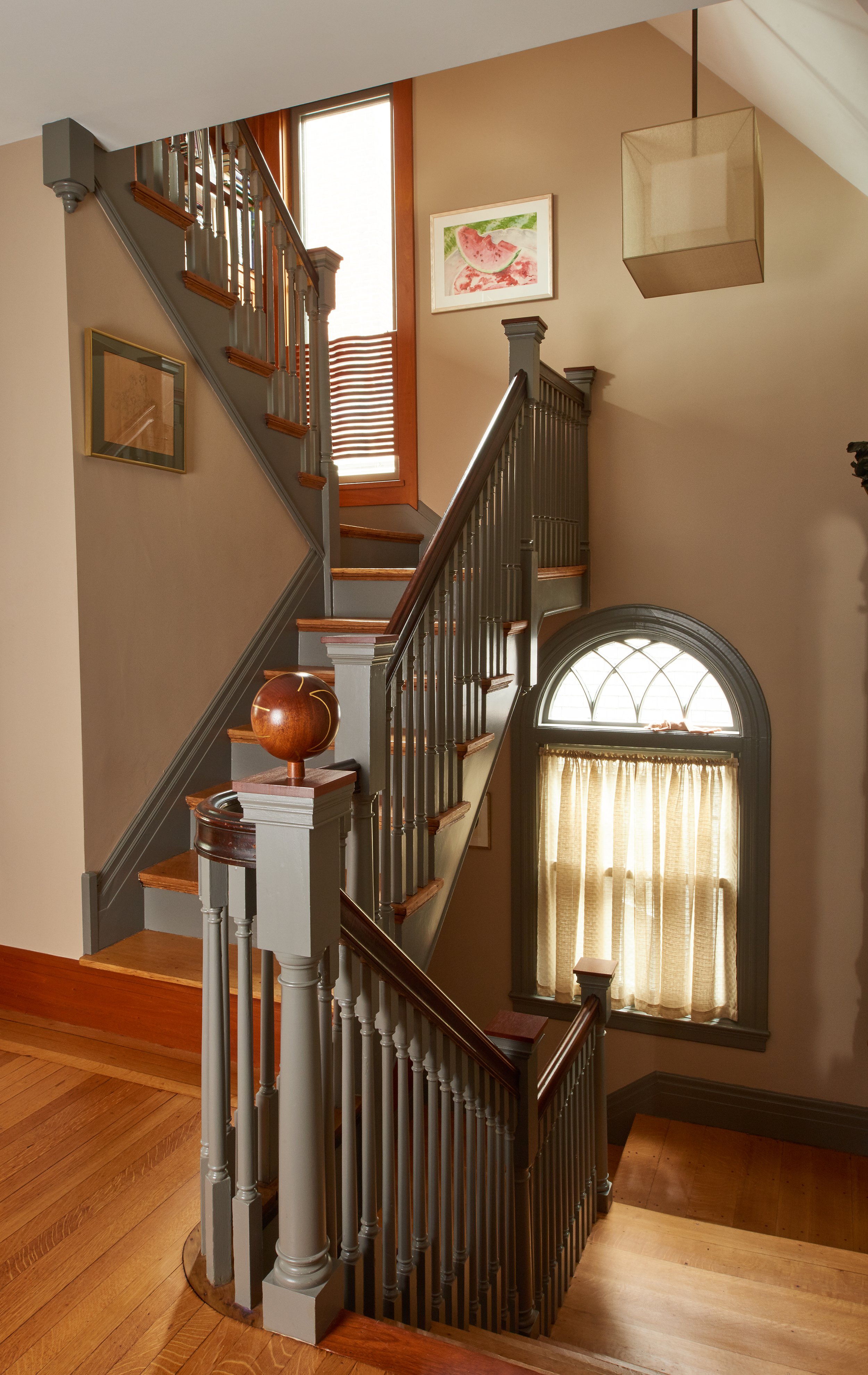 Interior view of a staircase with gray painted wooden balustrades, wooden handrails, and stairs, next to a large window with a curtain. There are wall art pieces, a decorative globe, and a modern pendant light.