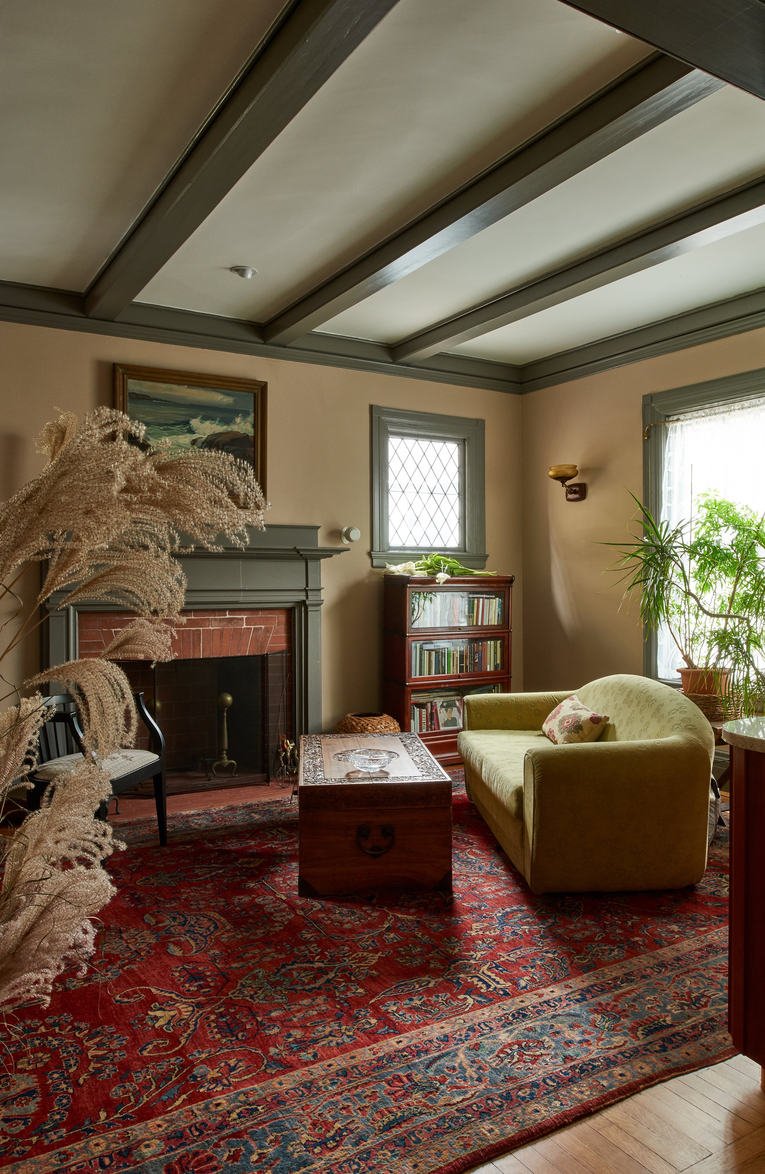 A cozy living room with a fireplace, a green armchair, a wooden coffee table, and a vintage red rug. There are bookshelves, potted plants, and a window with lace curtains allowing natural light in.