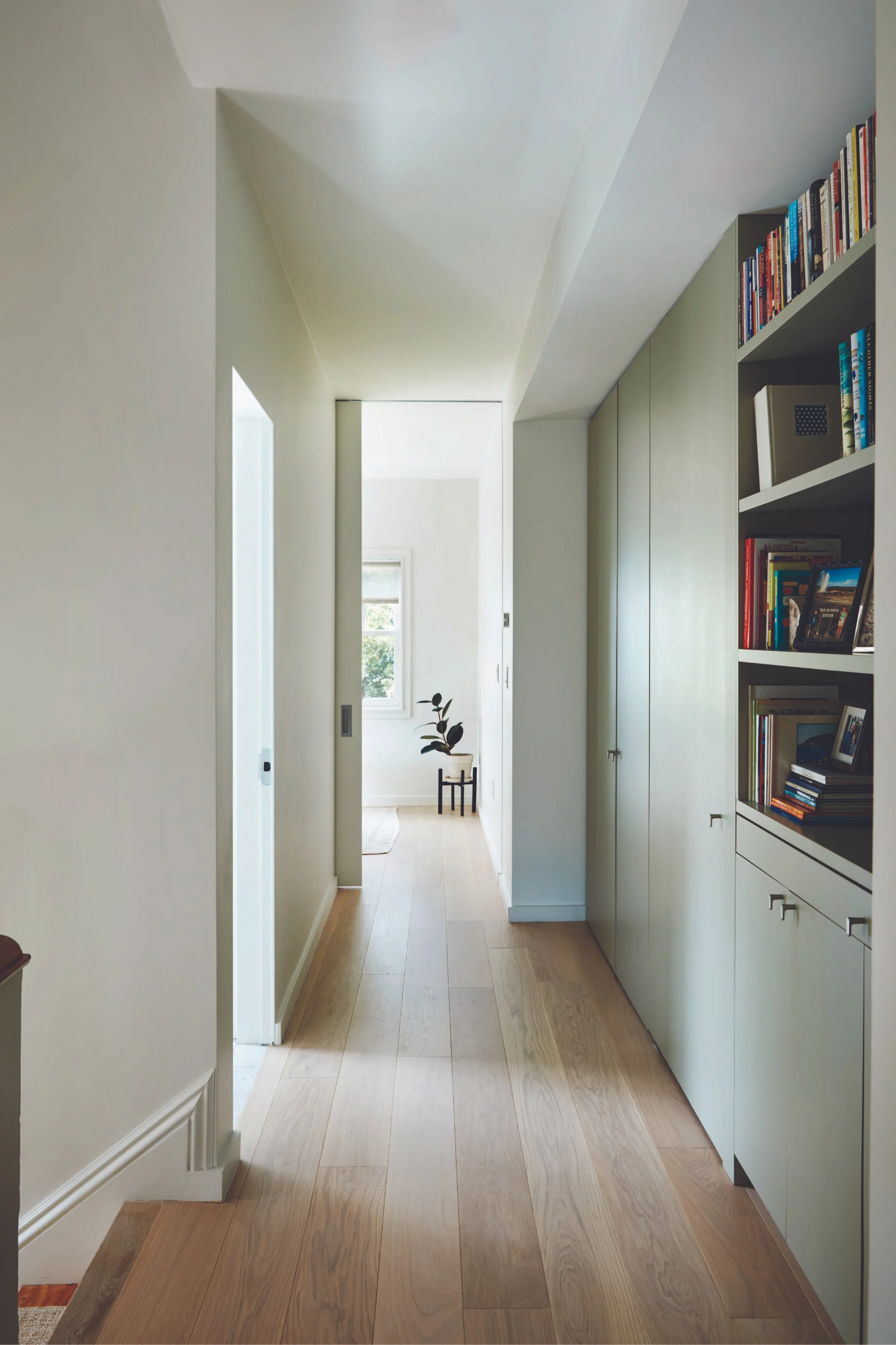 A hallway with light-colored wooden floors, white walls, and a built-in bookshelf filled with books on the right side. There is a small plant on a stand at the end of the hallway, near a window with sunlight streaming in.