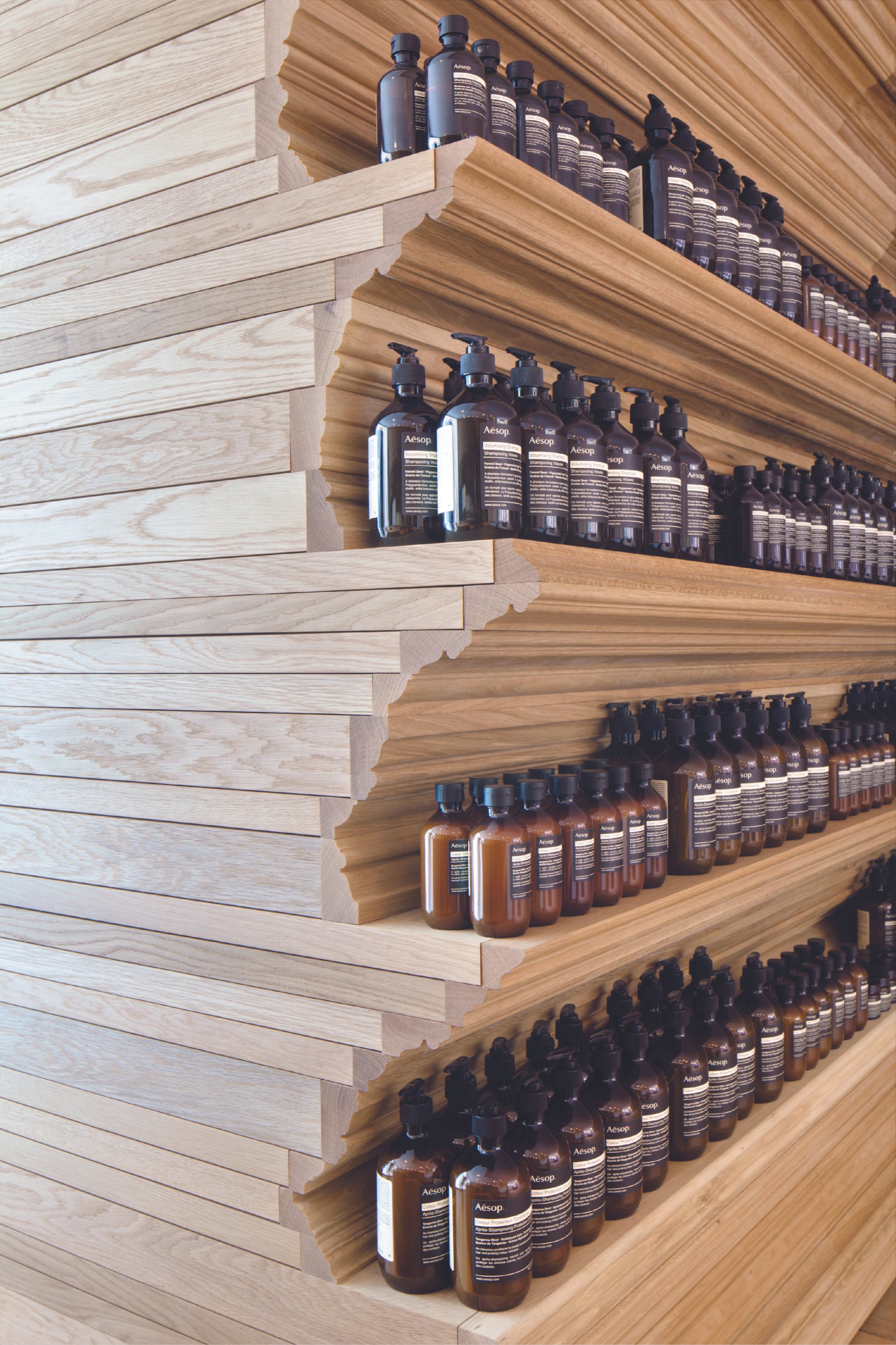 Wooden shelves displaying rows of brown bottles with black caps, labeled Aesop, inside a retail store.