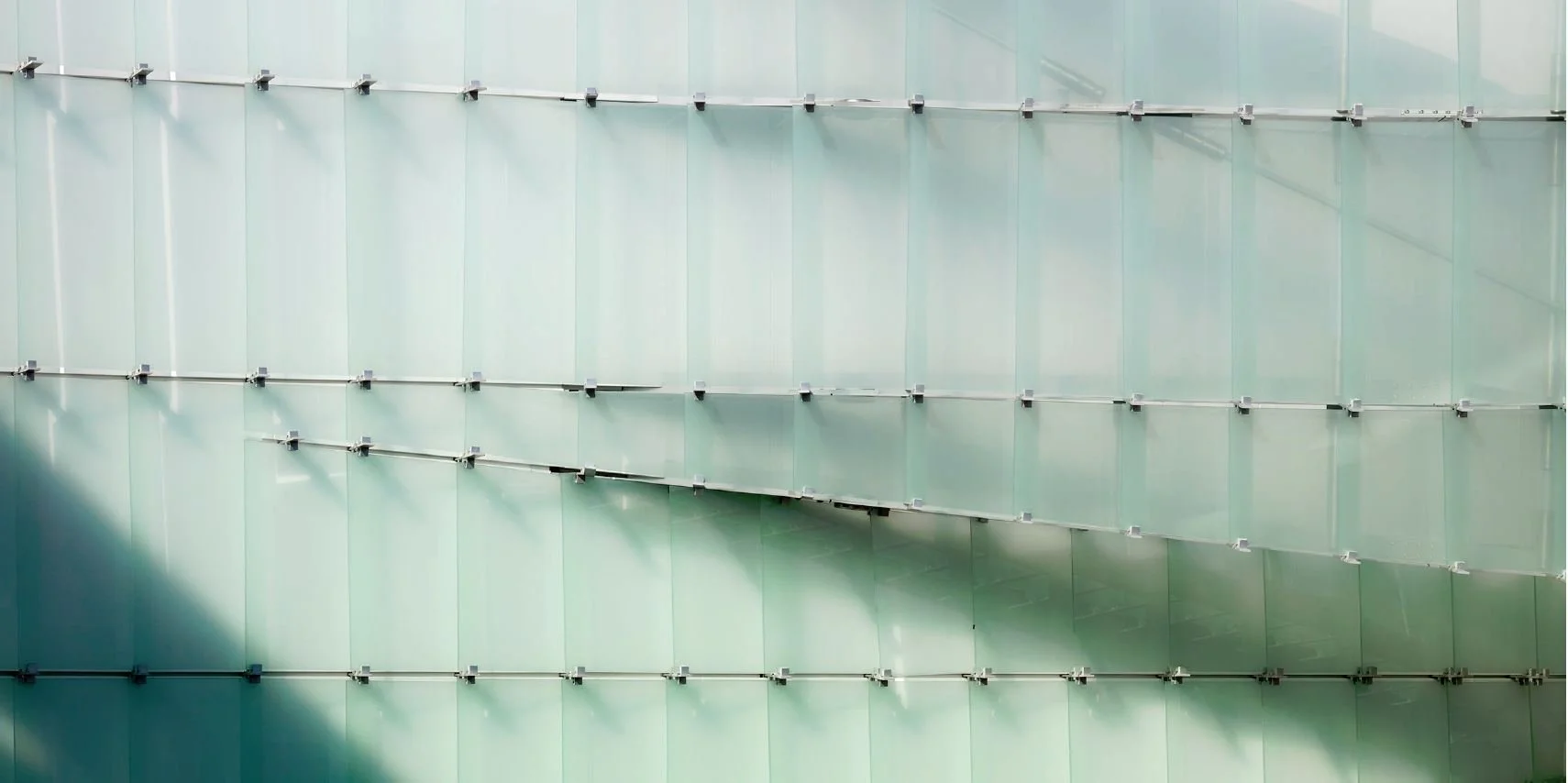 This image shows a modern architectural glass building facade with a grid of frosted glass panels held by metal brackets, reflecting sunlight and casting shadows.
